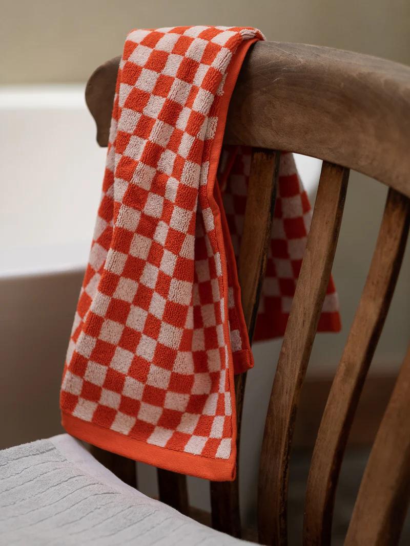 An orange and white checkered towel hangs over a wooden chair, alongside a Baina hand towel. A bathtub and gray bathmat are visible in the background.