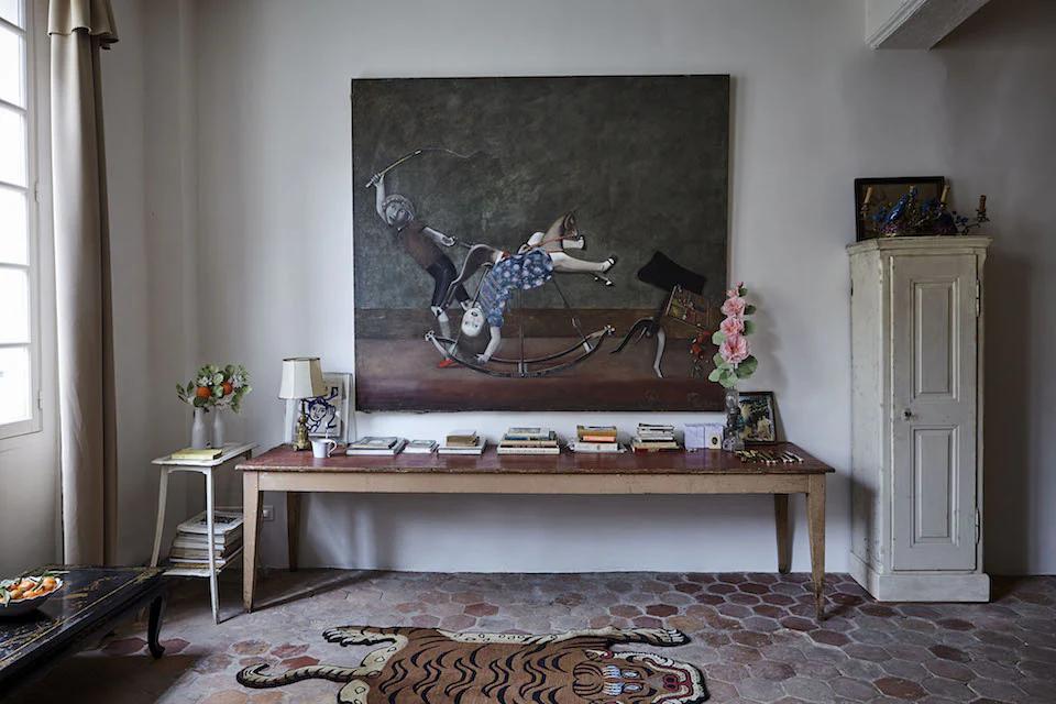 Parisian-style room with surreal painting above a wooden table. Books, lamp, and Astier de Villatte ceramics adorn the table, with a tall cabinet nearby.