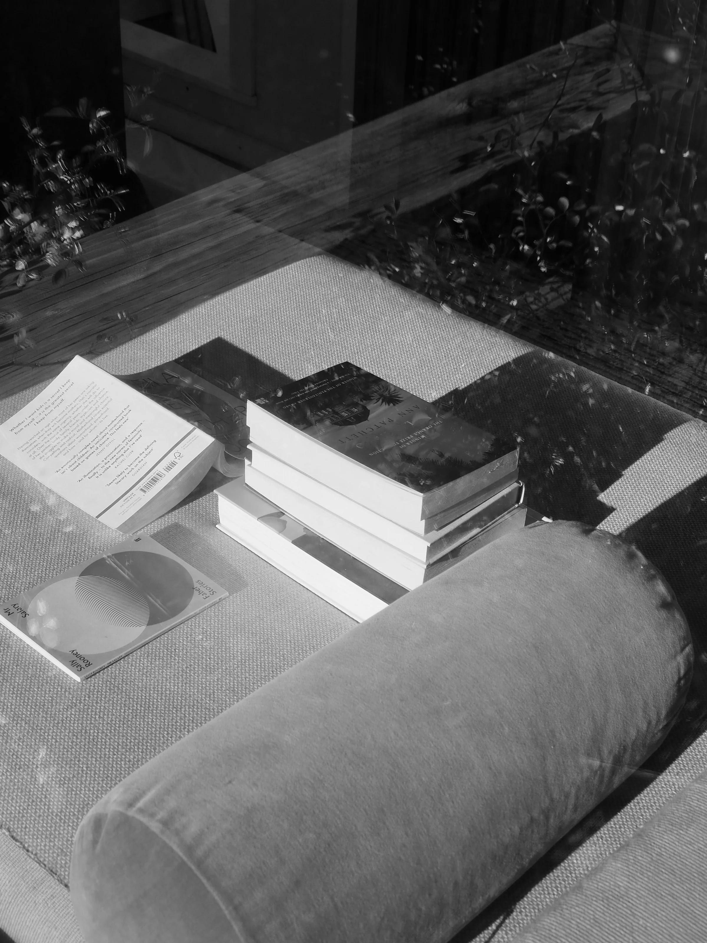 A black-and-white photo of books, a CD, and a long cushion on a sunlit window seat, reflecting outdoor trees and creating a peaceful, reflective mood.