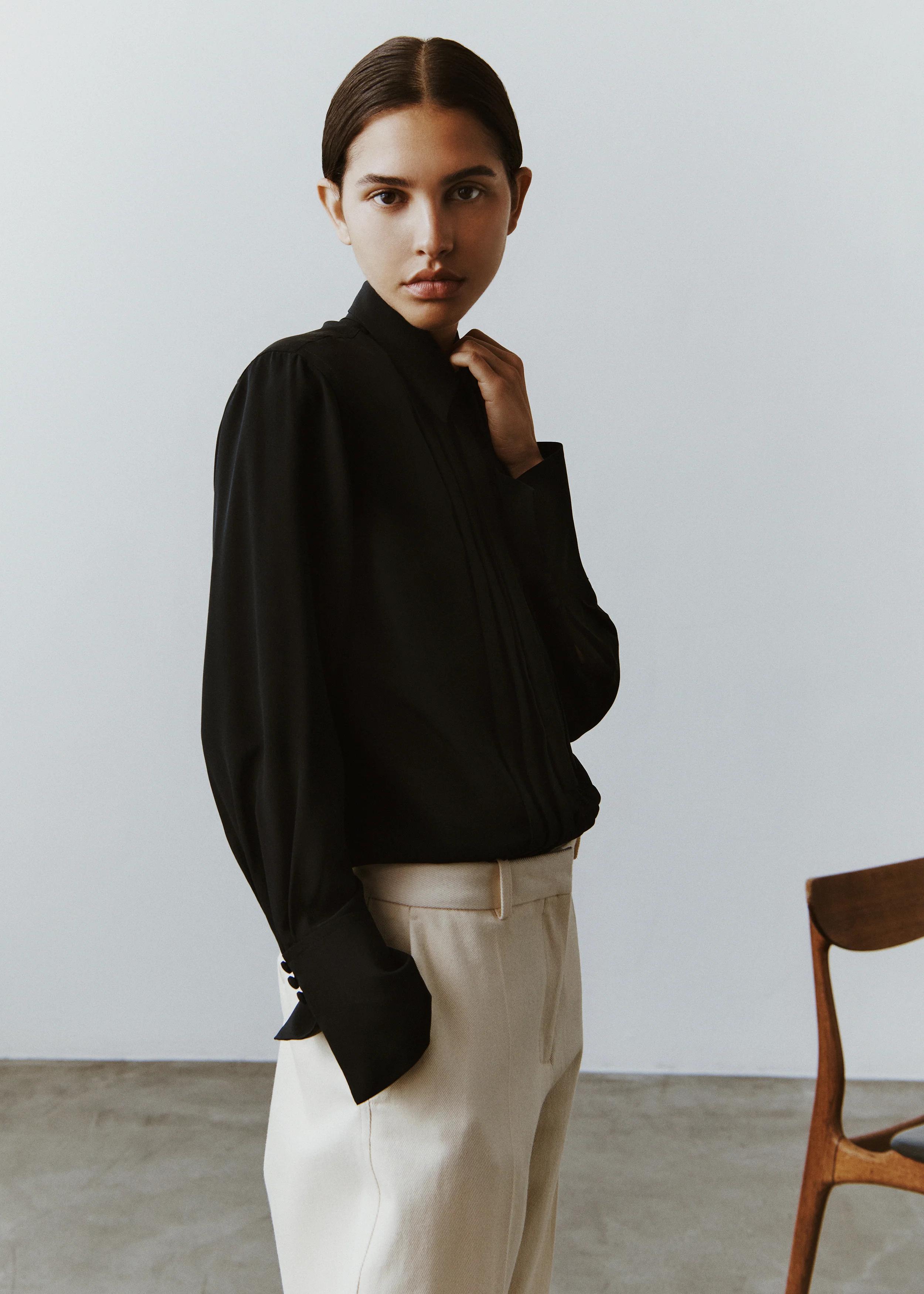 Person with dark hair in black long-sleeve blouse and beige pants. Standing in light-coloured room, touching collar. Wooden chair visible.