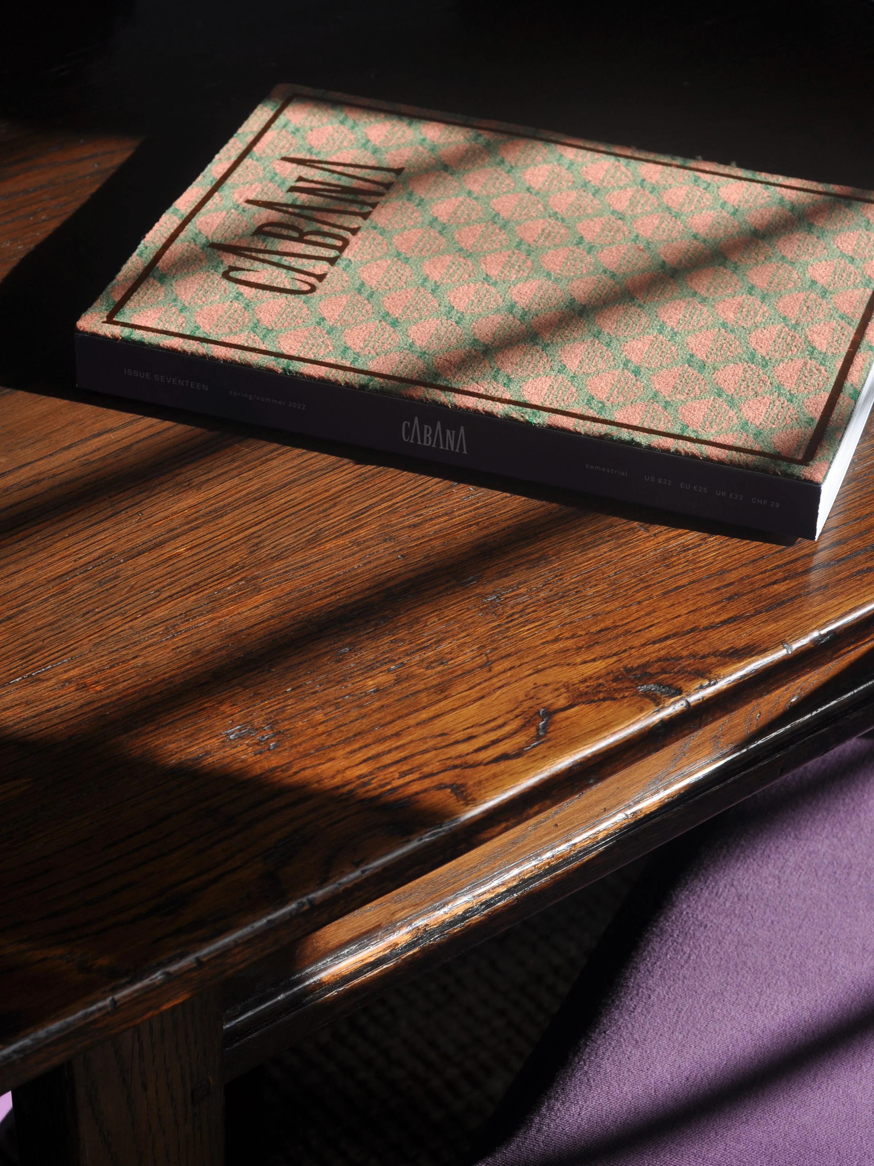 "Cabana" book with textured cover on wooden table. Diagonal sunlight casting shadows. Purple surface edge visible.