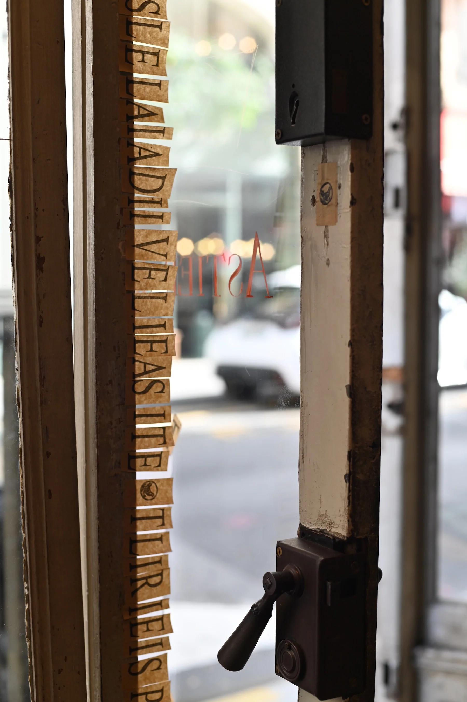 Close-up of old wooden door with metal latch. Peeling gold letters on glass pane. Blurred street scene outside.