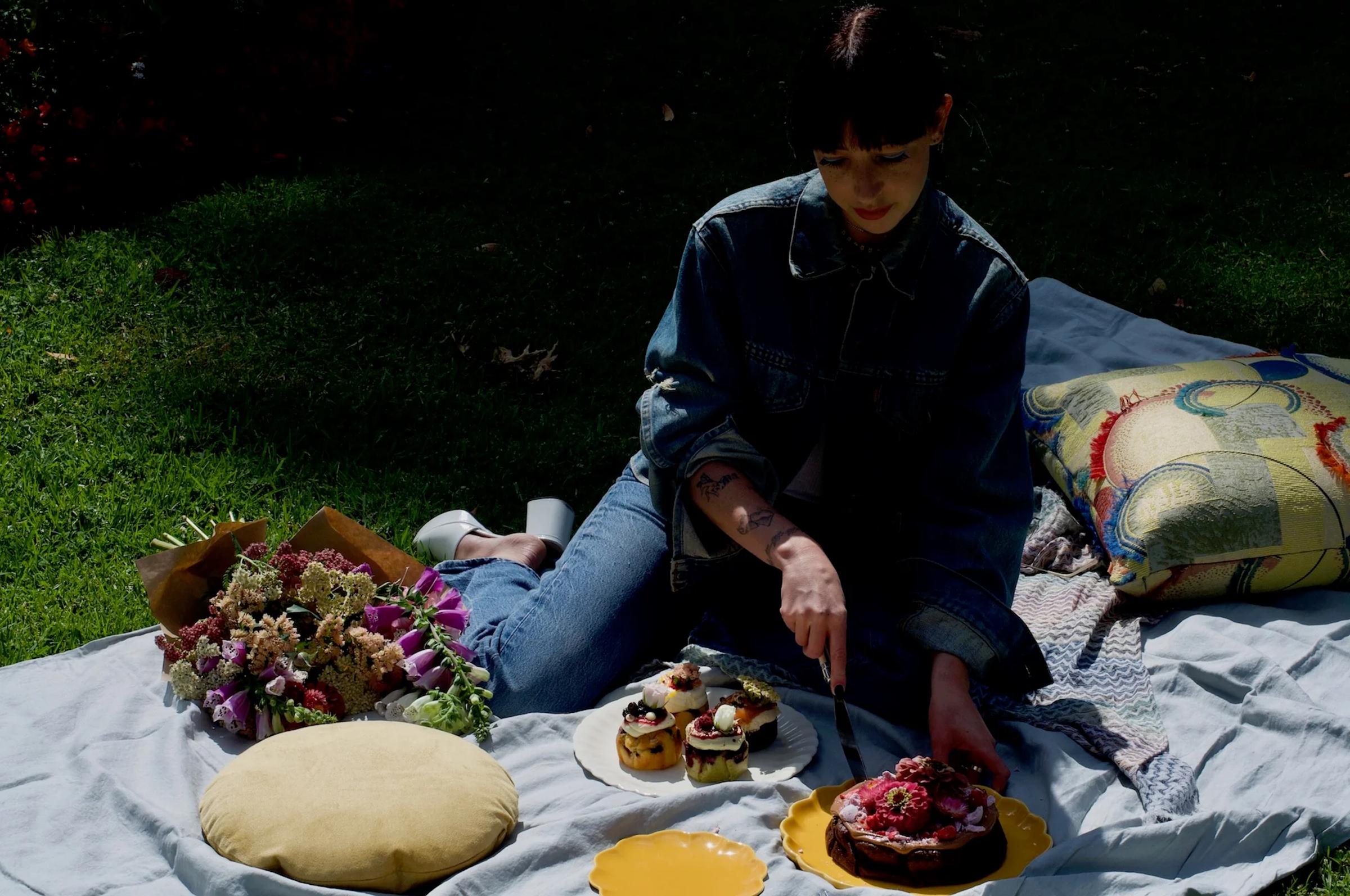 A person in a denim jacket slices a cake from The Caker on a yellow plate, with flowers and pastries nearby on a shaded picnic blanket.