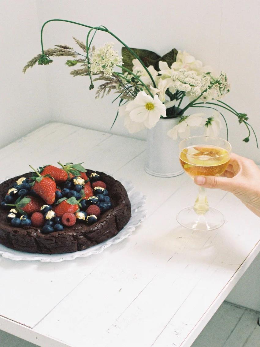 A chocolate cake topped with berries and edible gold flakes on a white plate, with a hand holding a glass of wine and a vase of flowers in the background.