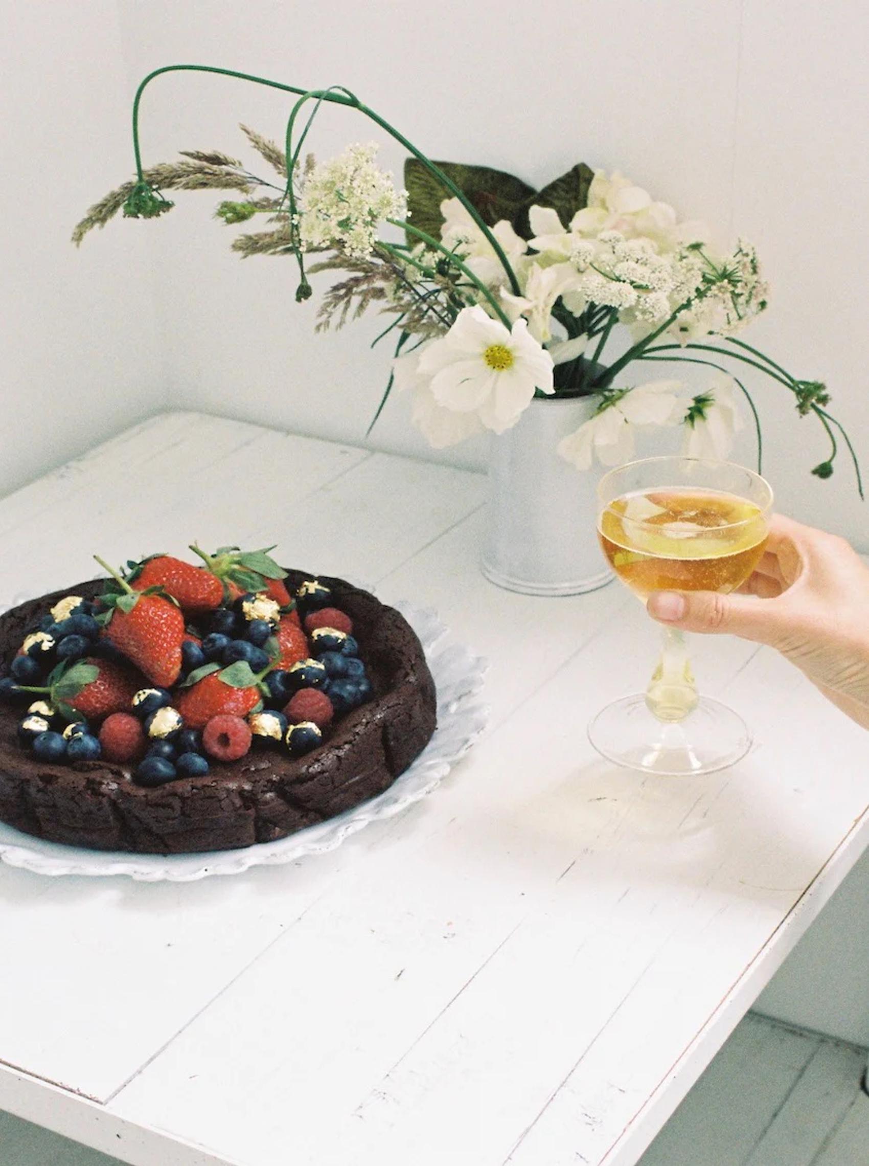 A chocolate cake topped with berries and edible gold flakes on a white plate, with a hand holding a glass of wine and a vase of flowers in the background.