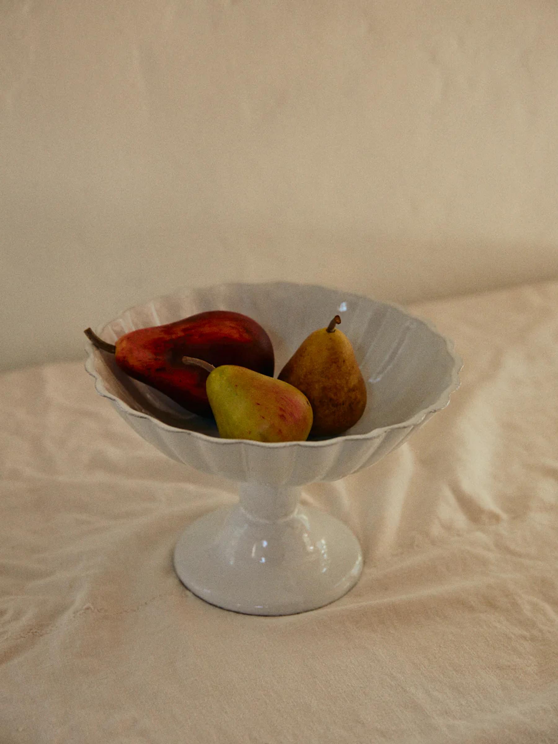 White pedestal bowl with four carved pears, two yellow and two red, on beige tablecloth. Soft lighting casts gentle shadows.