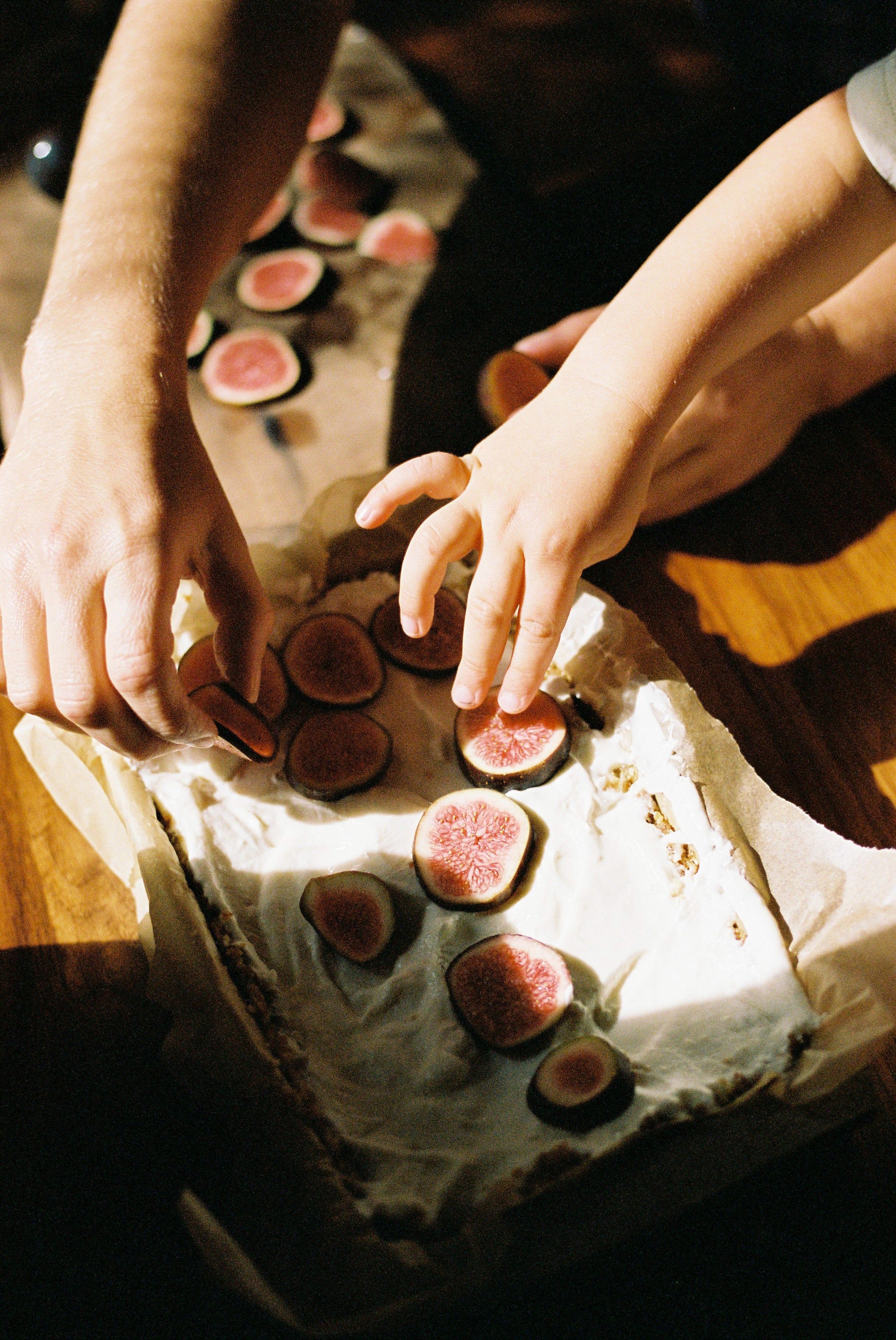 Adult and child hands arranging fig slices on cream-covered tray. Warm, natural lighting.