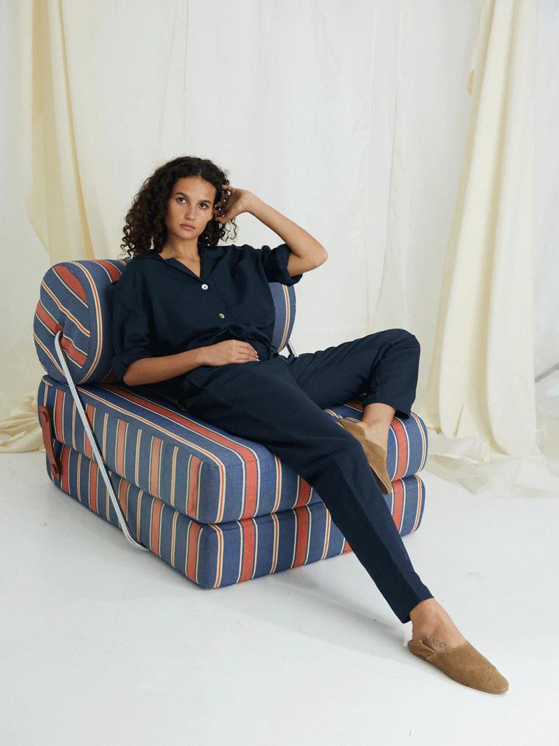 Person in stylish sleepwear sits on striped chair. Curly hair, brown slippers. Hand rests on face. Light fabric background.