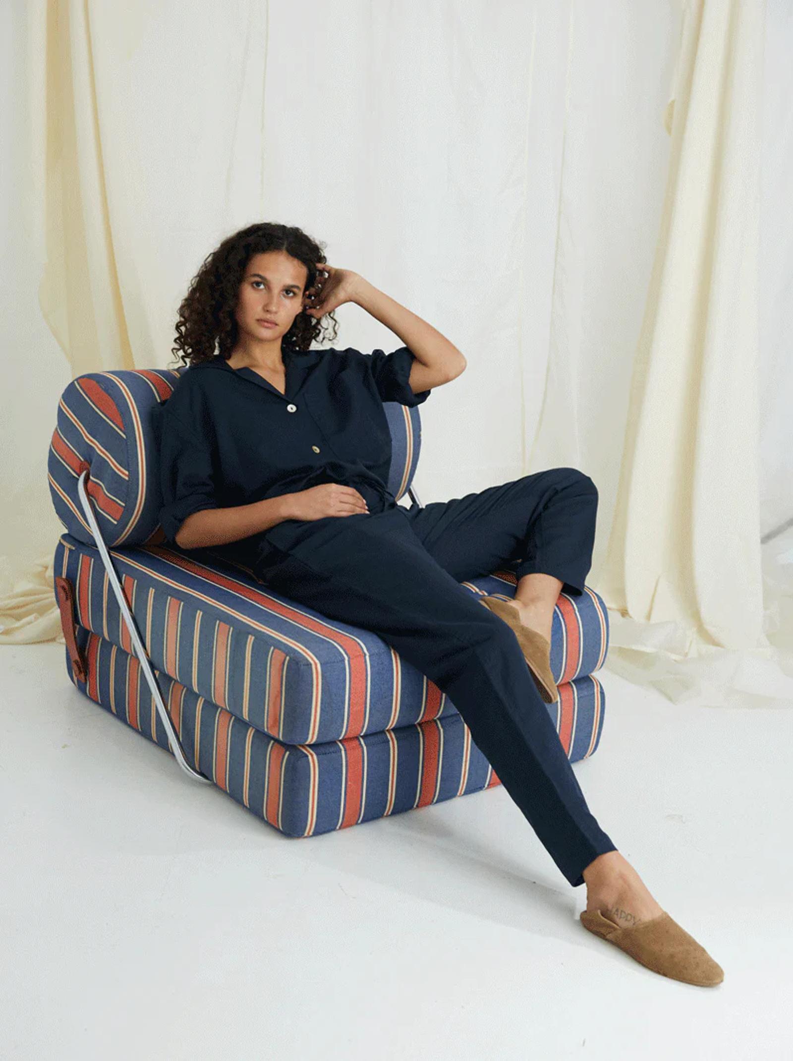- Person in stylish sleepwear sits on striped chair. Curly hair, brown slippers. Hand rests on face. Light fabric background.