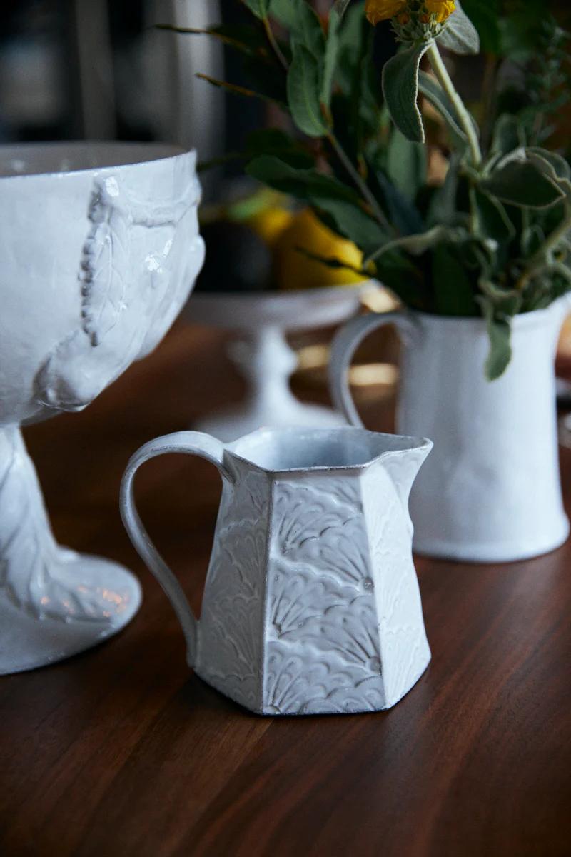 White floral pitcher on wooden table. Similar vase with greenery and yellow flowers in background. Evokes warmth of Italian dining.