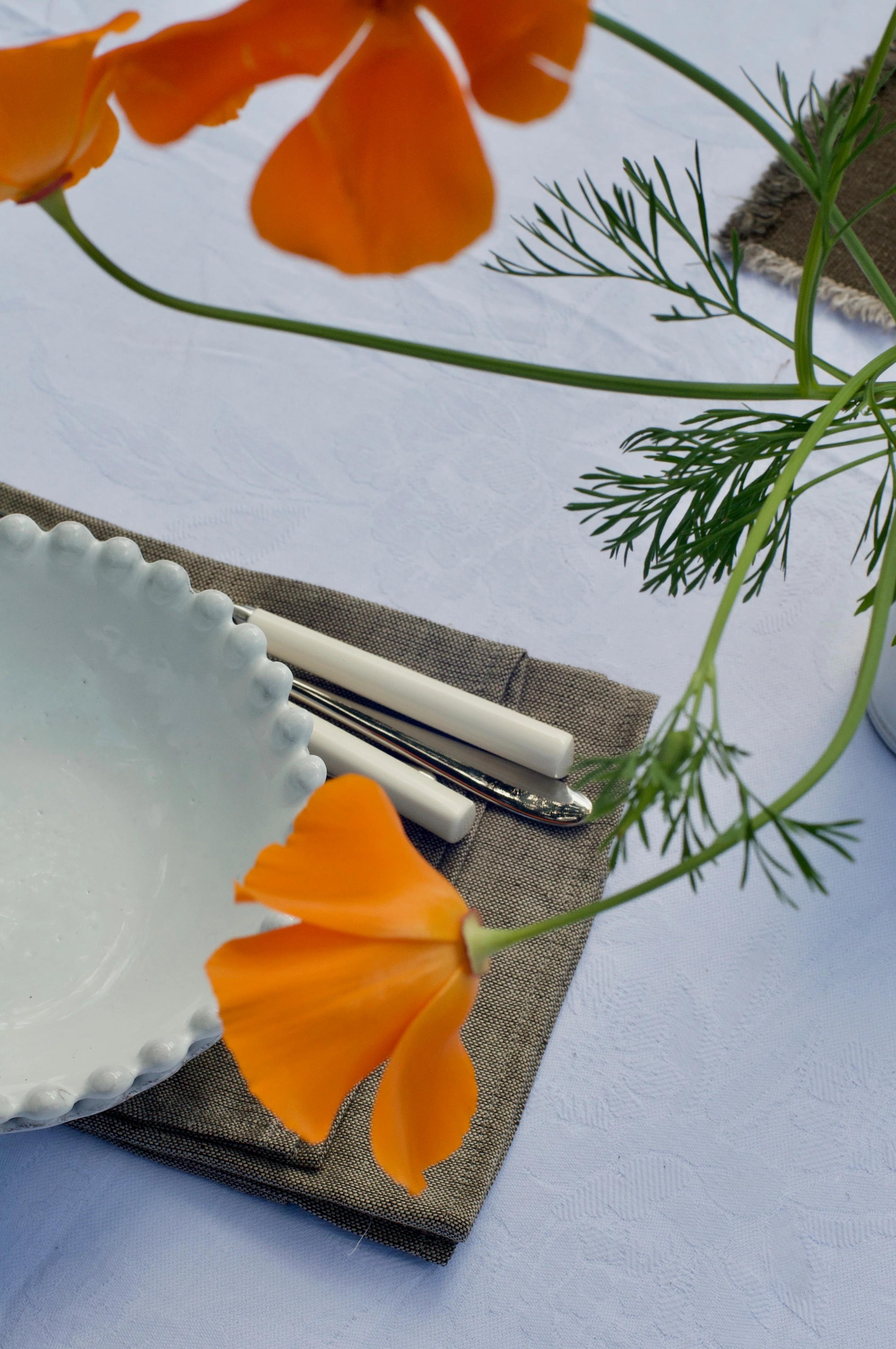 White scalloped bowl on taupe napkin. White-handled cutlery nearby. Orange flowers add vibrancy to contemporary design.