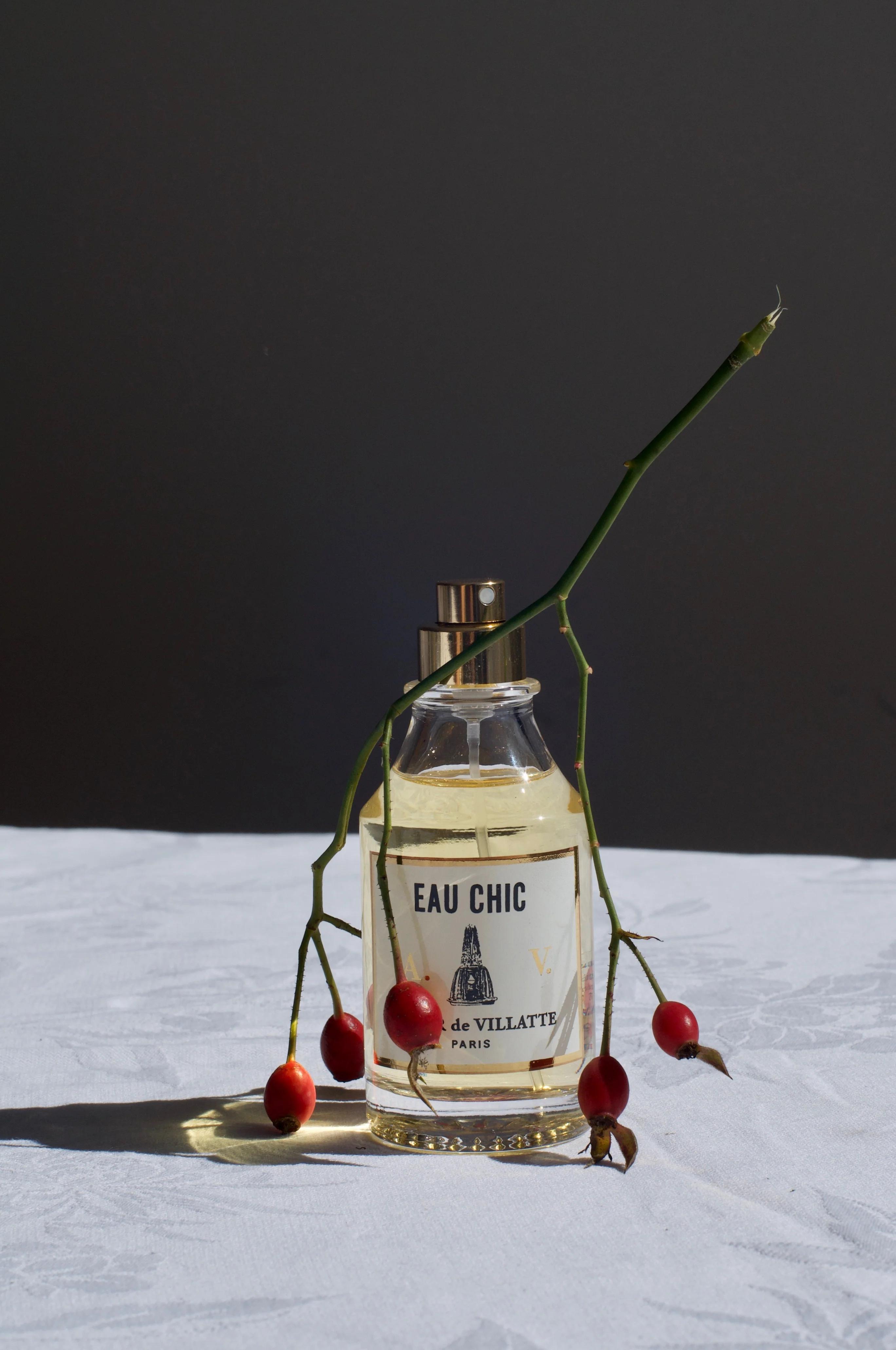 Eau Chic de Villatte Paris perfume bottle on a white cloth, adorned with an upside-down sprig of berries. The neutral dark background highlights the golden hue of the cologne.