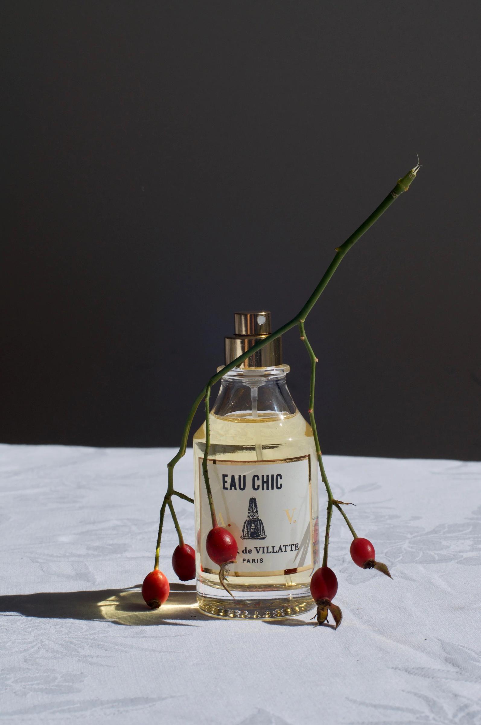 Eau Chic de Villatte Paris perfume bottle on a white cloth, adorned with an upside-down sprig of berries. The neutral dark background highlights the golden hue of the cologne.