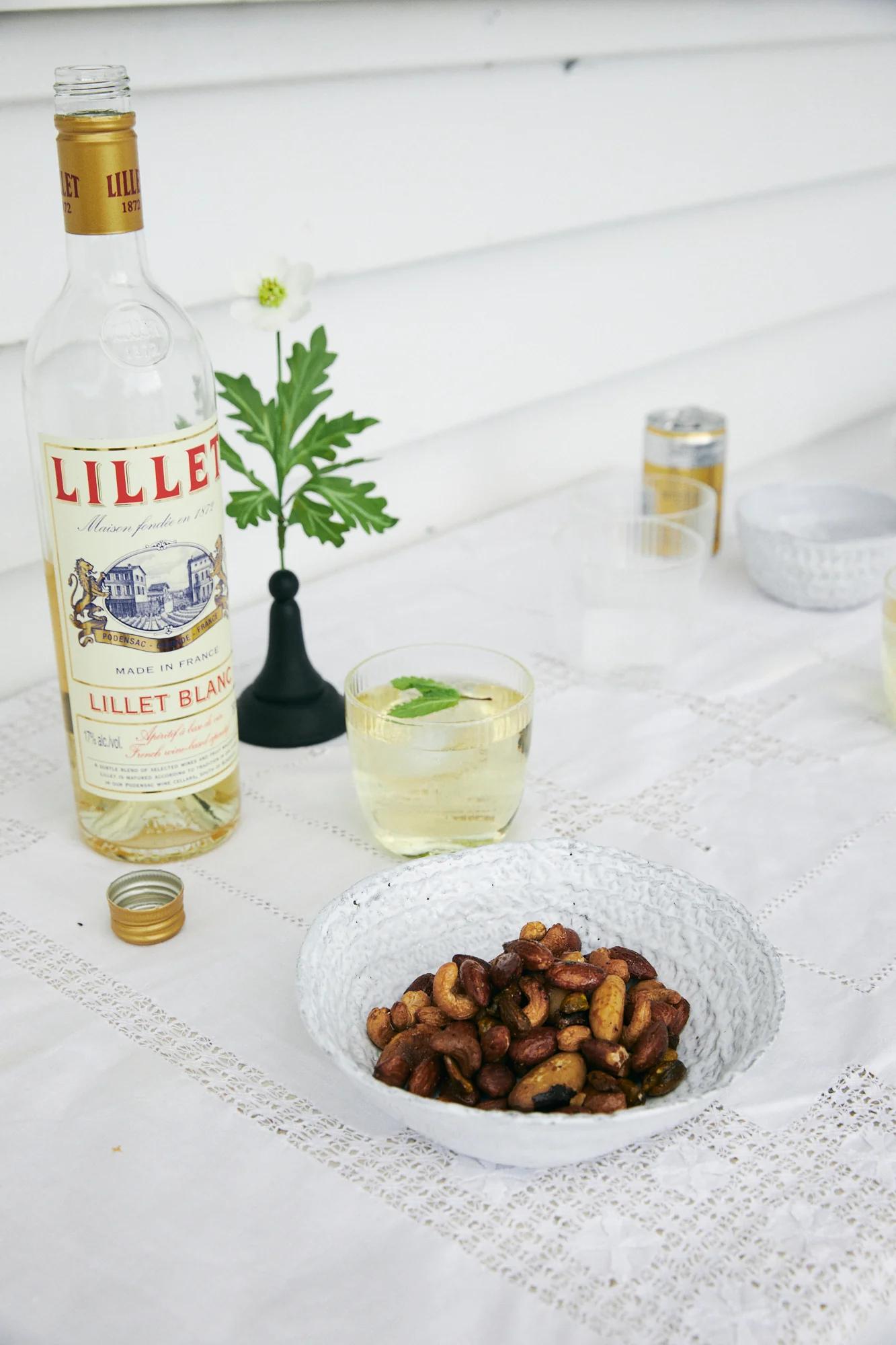 Lillet Blanc bottle, small black vase with flower, glass of light drink, and bowl of nuts on white tablecloth.