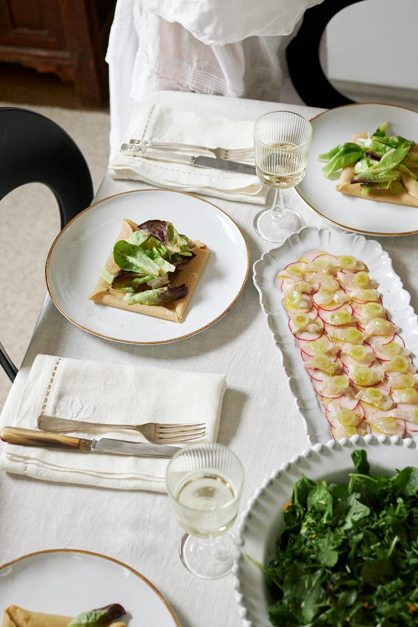 A festive table with white linens and plates of savoury dishes topped with greens. Radishes with fish, salad, and wine glasses evoke South of France dining.