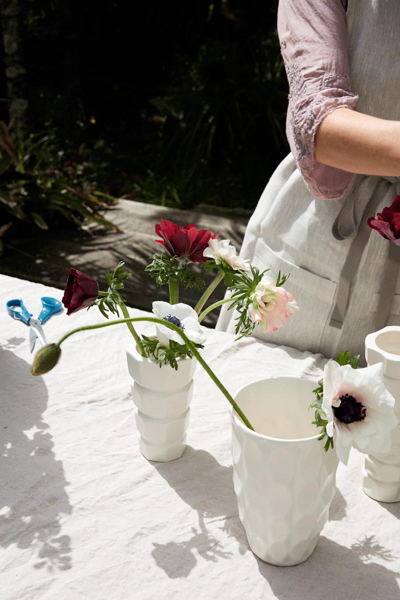 Person in a pink shirt and grey apron, arranges flowers at an outdoor table. Three white vases hold red, white, and purple flowers, creating a warm, inviting dinner party scene.