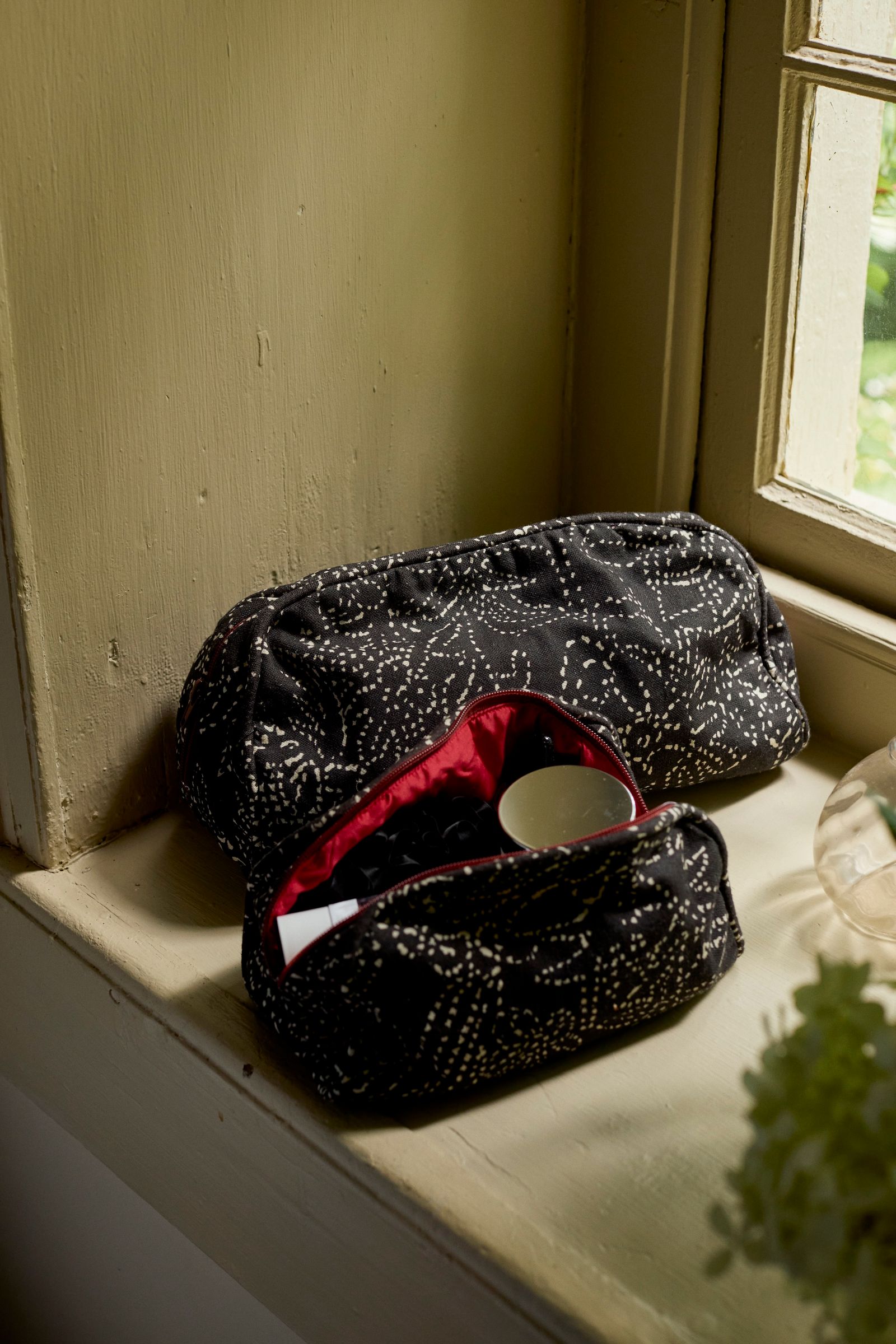 two toiletry bags sit on a green window sill, one opened to reveal cosmetic jars