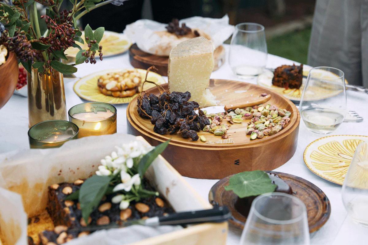 Outdoor table with cheese platter, desserts, flowers, candles, and wine glasses on white tablecloth with yellow plates.