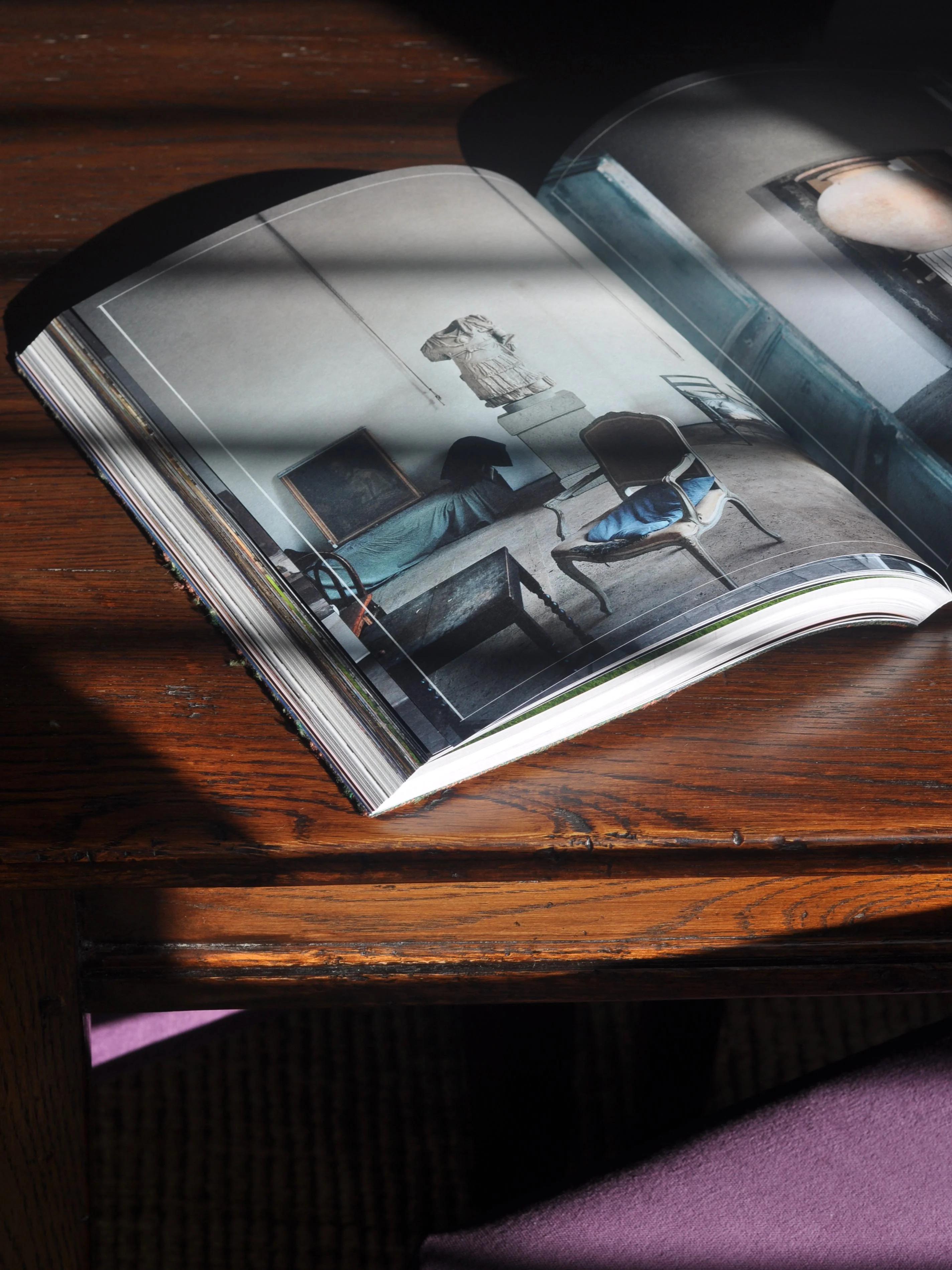 Open book on wooden table in natural light. Minimalist interior design photo visible. Shadows creating serene atmosphere.