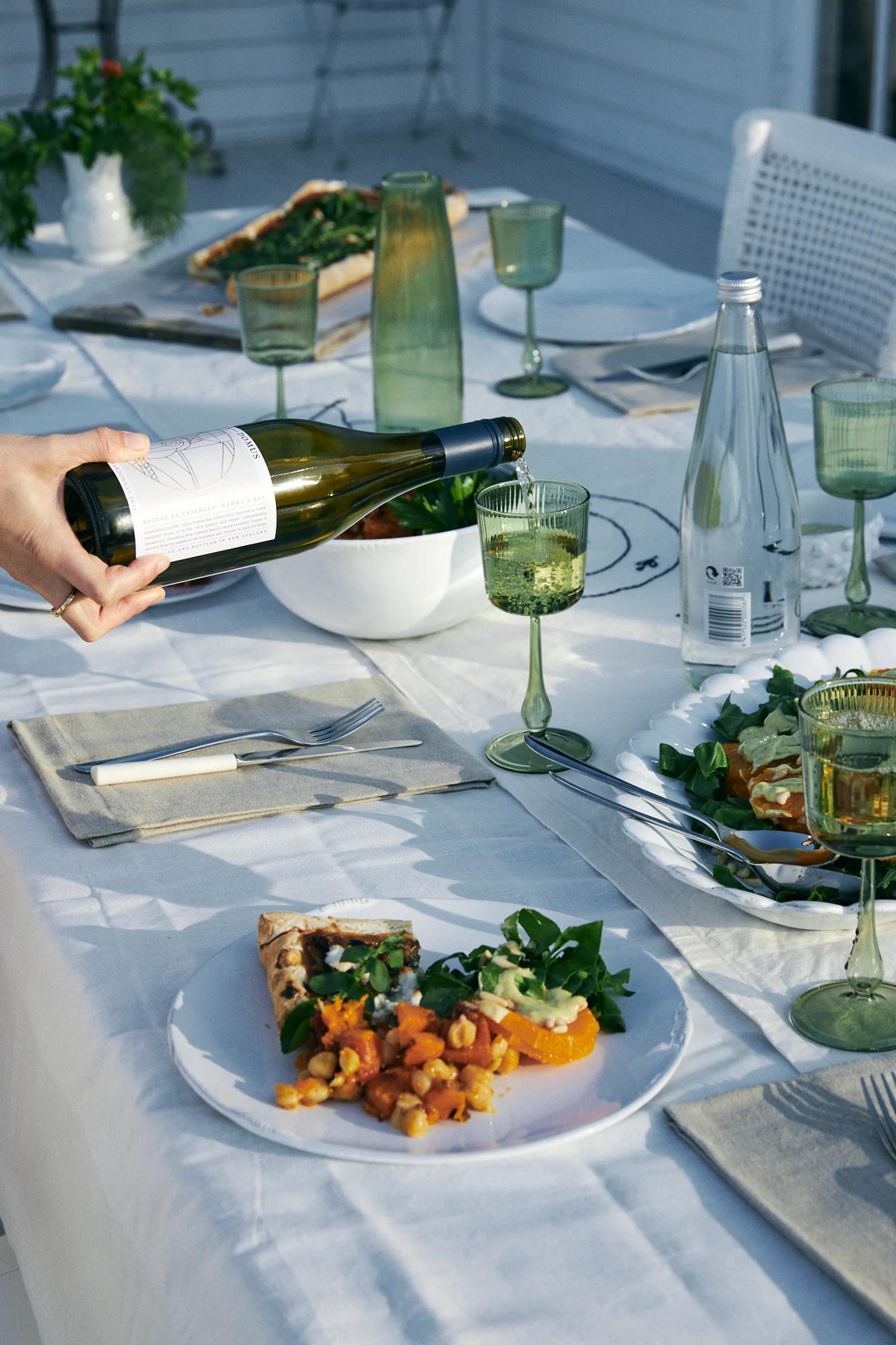 Outdoor table with pizza, salad, green glassware, wine being poured. White tablecloth, plants, bread visible.