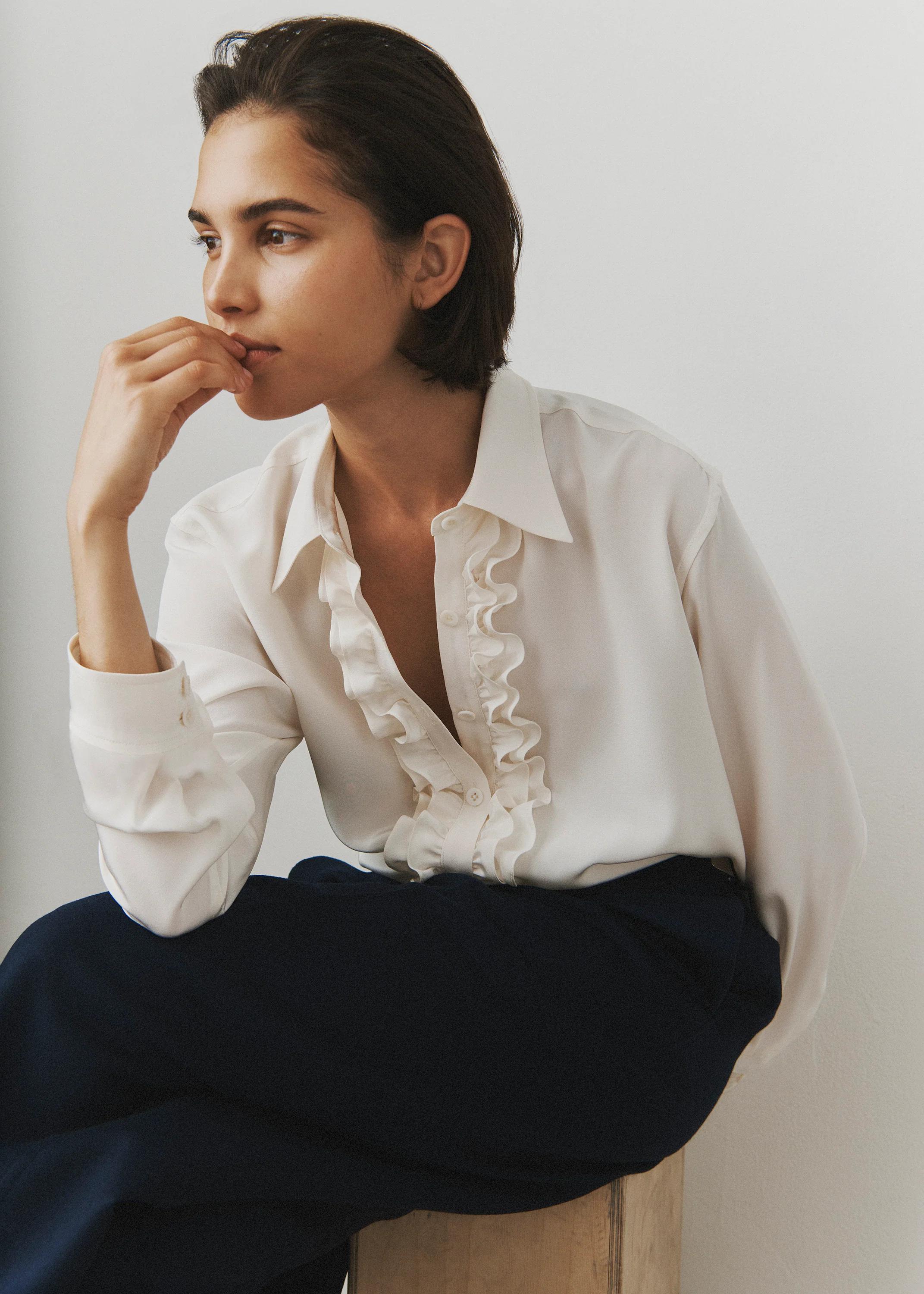 Person with short dark hair on wooden stool. White blouse with ruffled trim, dark pants. Hand on chin, looking left.