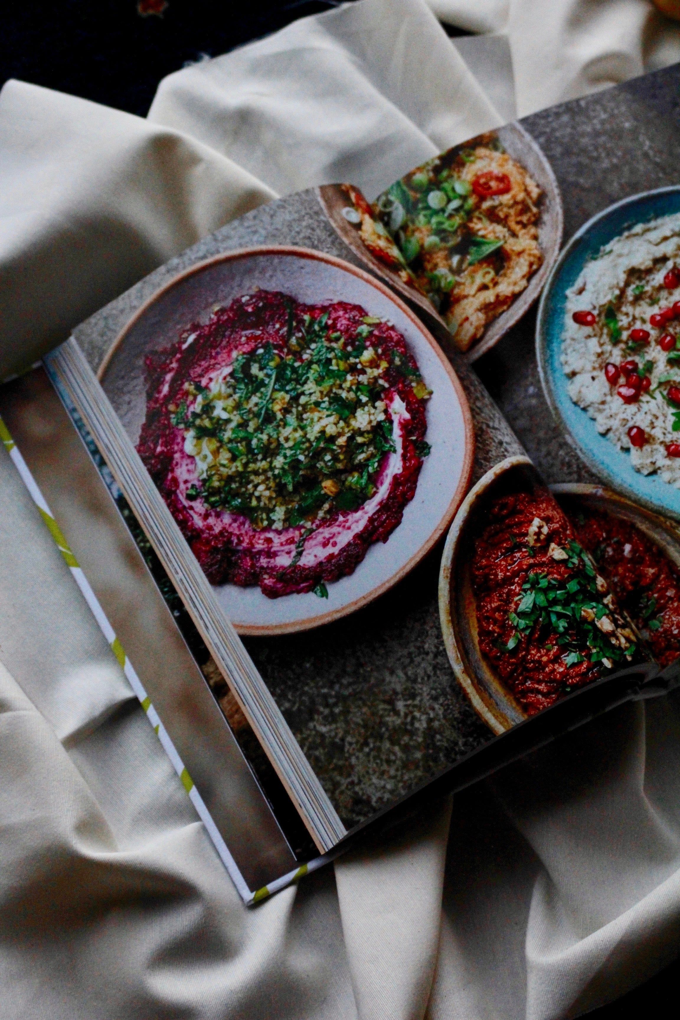 Falastin: A Cookbook_ lies open on a white cloth, displaying beetroot dip and creamy spreads garnished with herbs and colourful toppings.