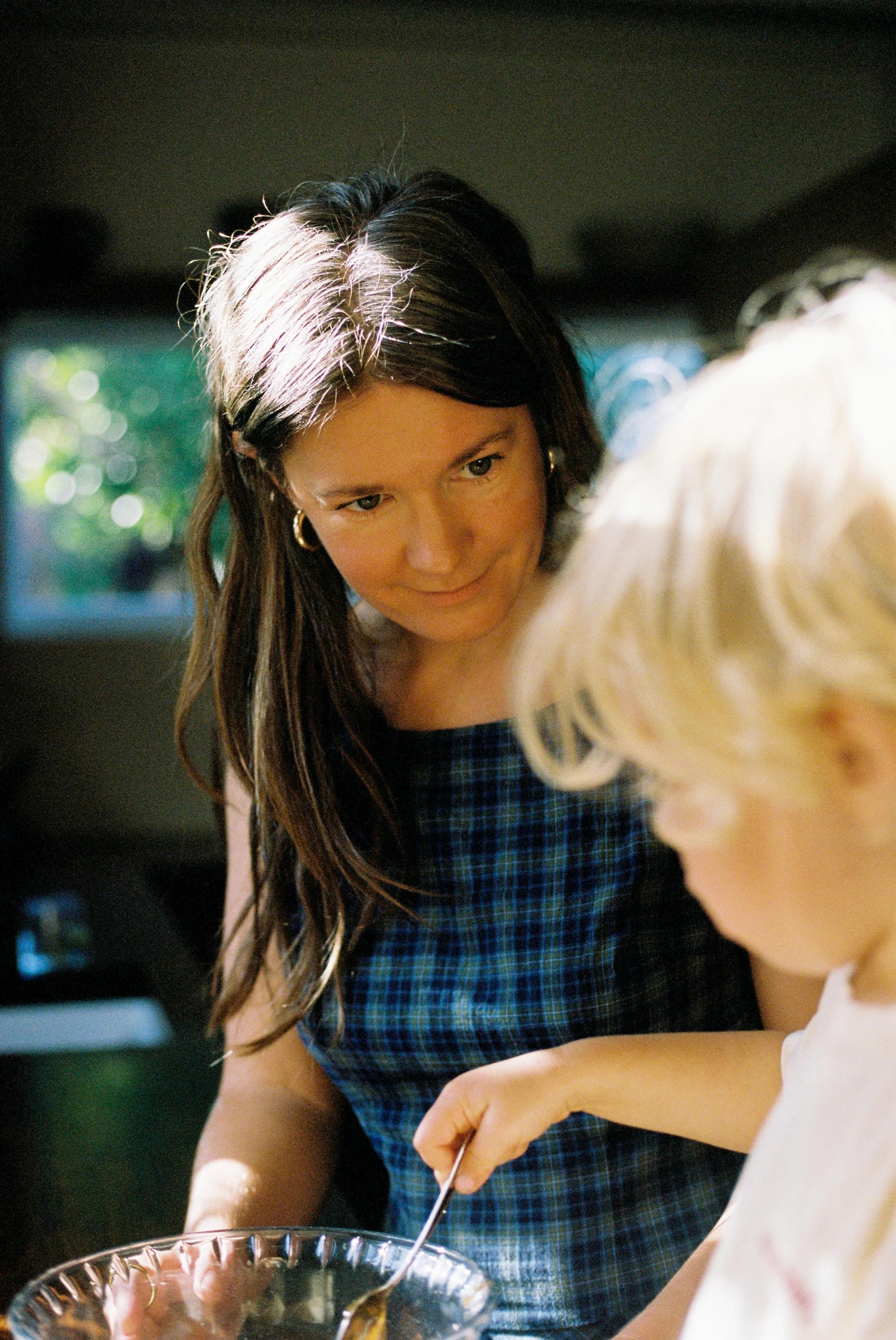 Woman in blue plaid dress looking at child stirring in glass bowl. Softly lit background.