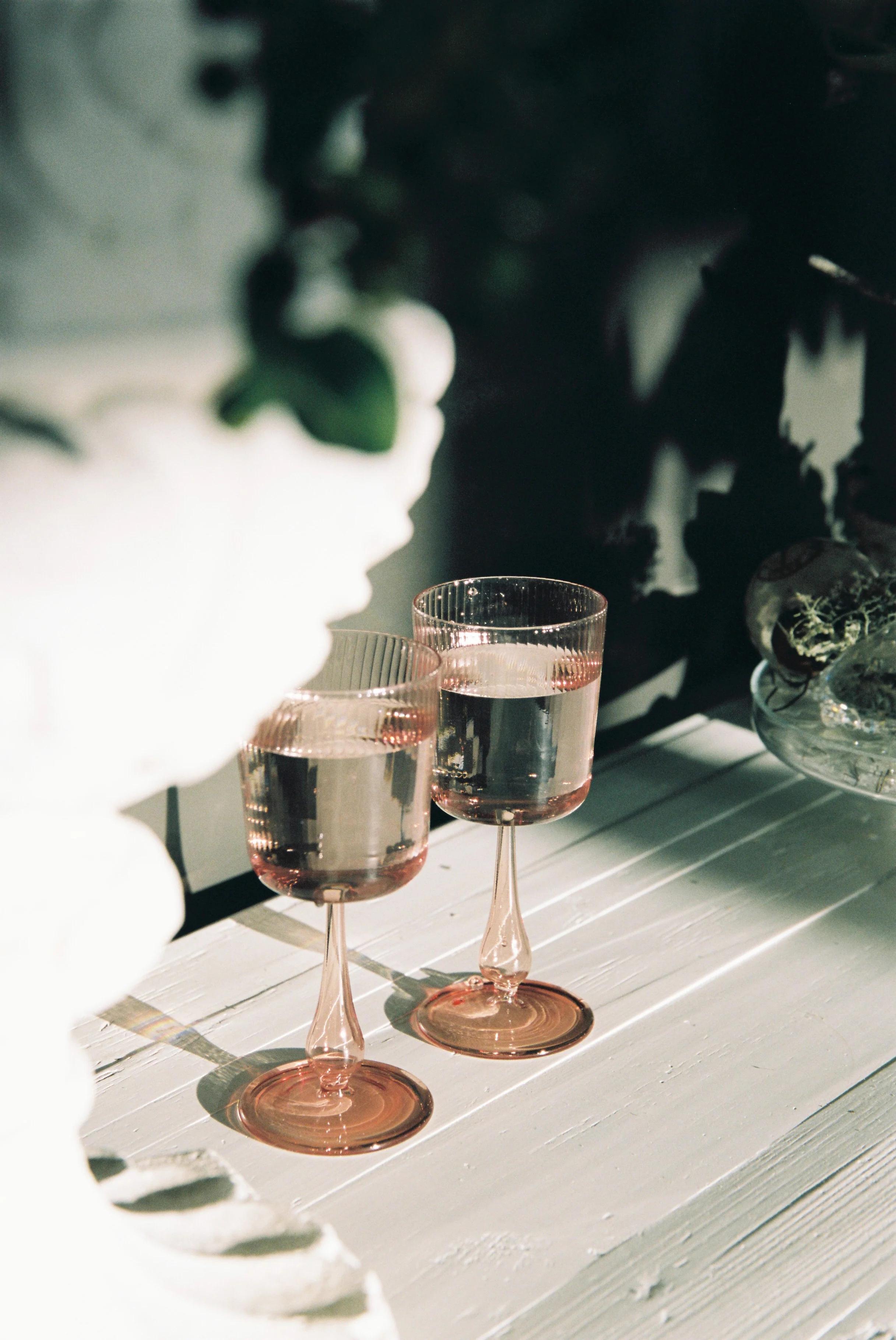 Two elegant R+D.LAB wine glasses filled with liquid sit on a white, rustic wooden table. Soft light and shadows create an intimate atmosphere against a dark, blurred background.