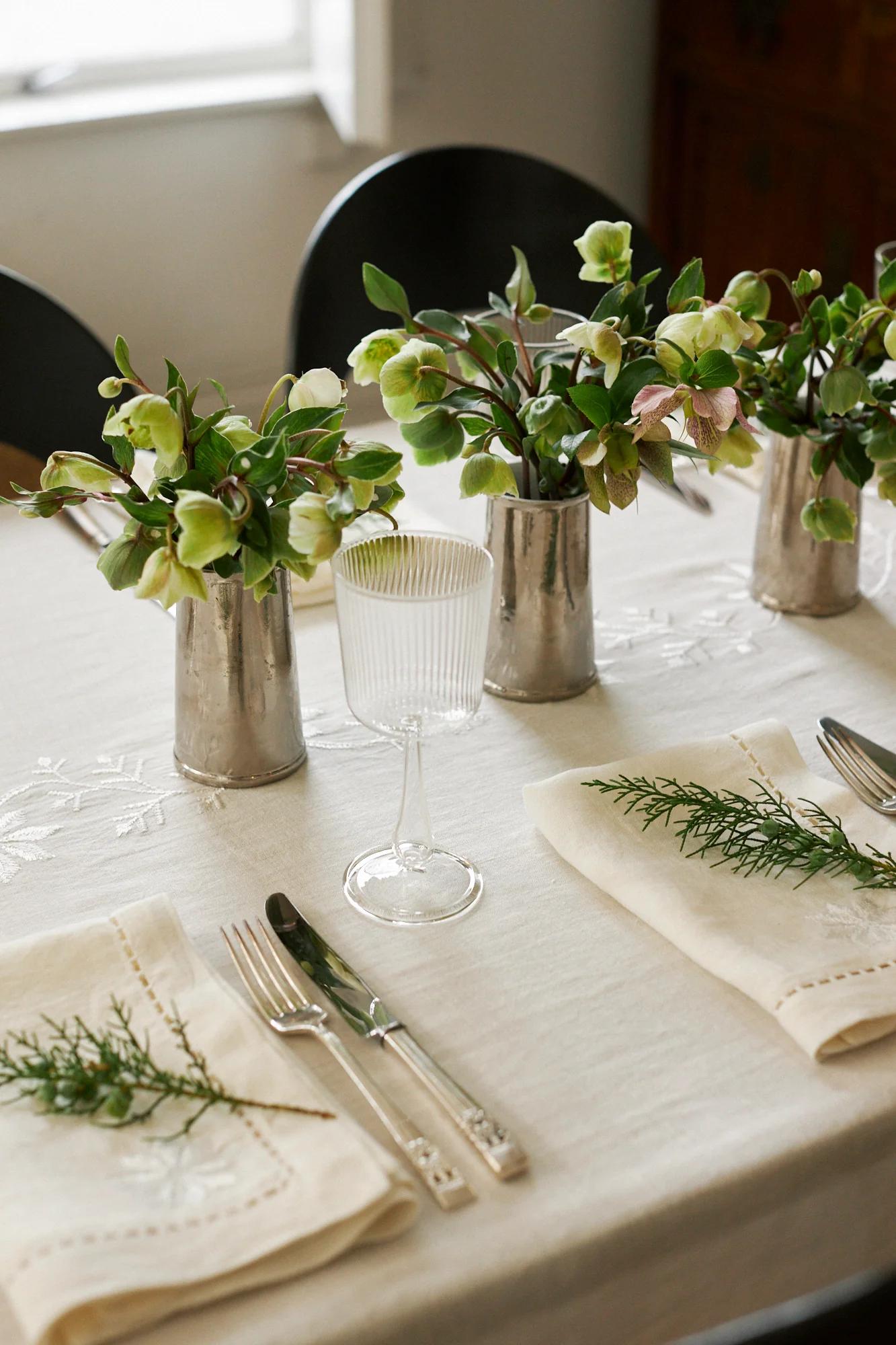 An elegant table with white linens, silverware, and napkins with greenery. Small vases of flowers and ribbed glasses complete the festive setting.