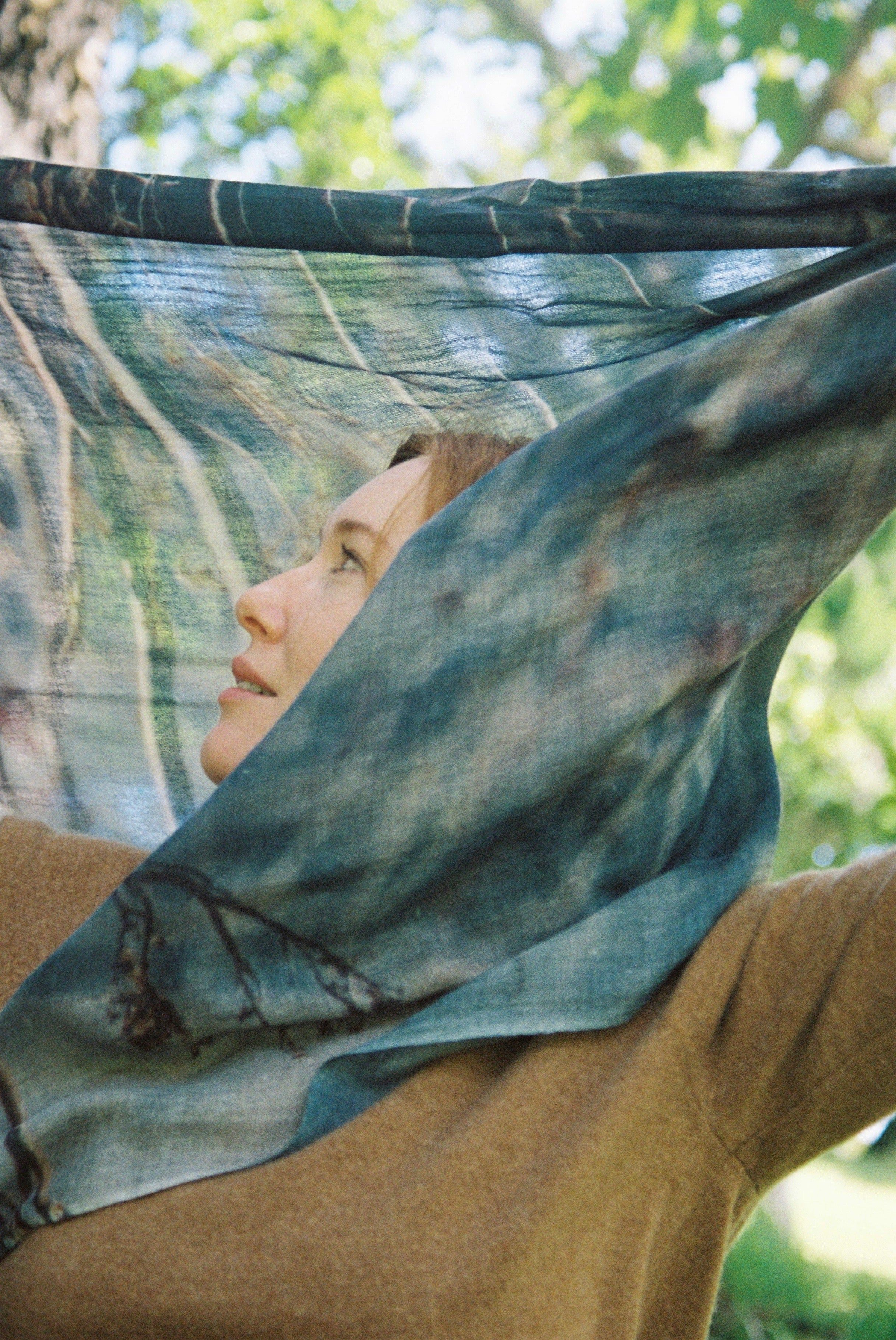 Person with light brown hair in brown top, wearing blue-green patterned scarf. Looking sideways against blurred green foliage background.