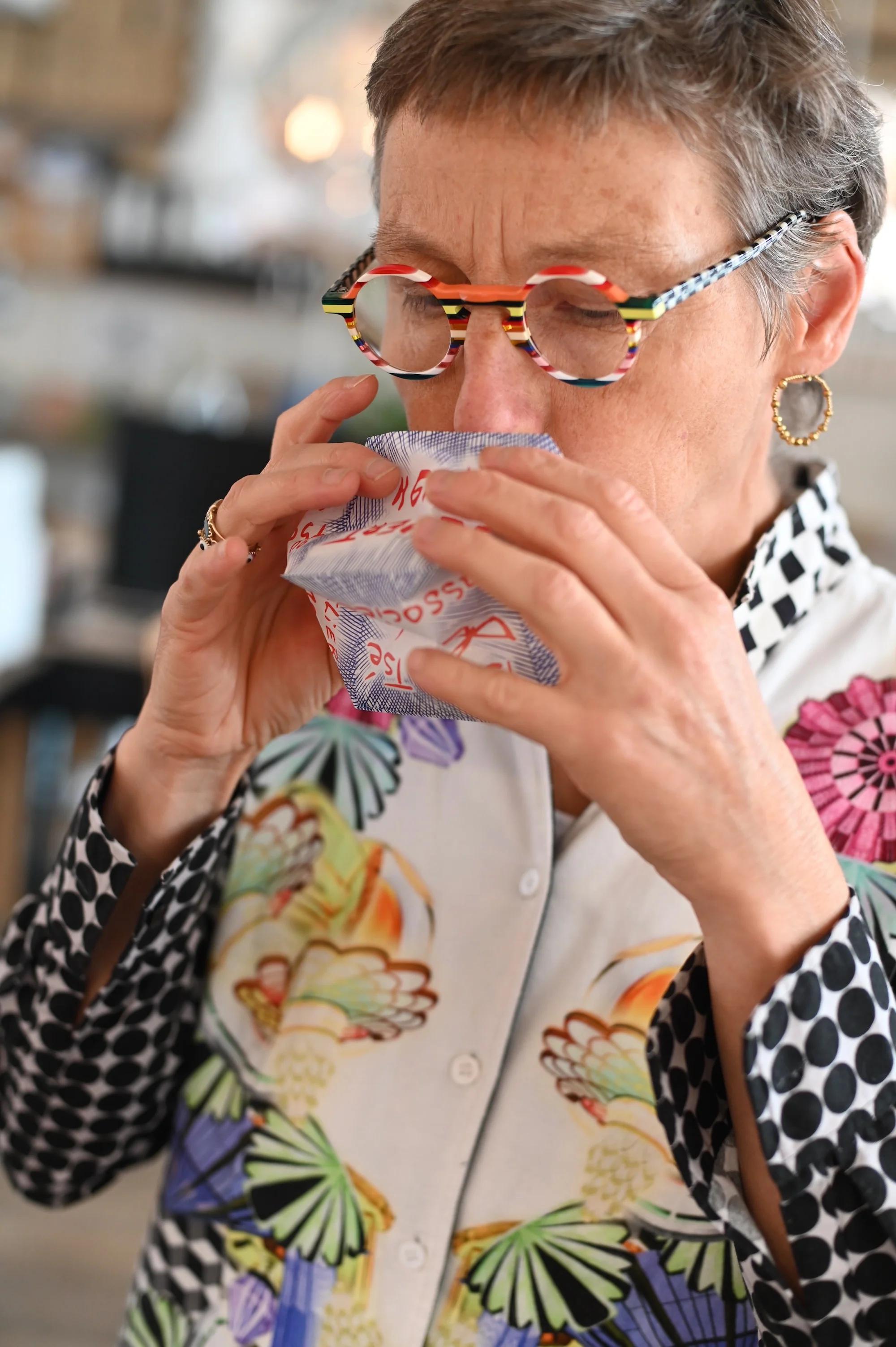 Person with grey hair, colourful glasses inspecting patterned cloth. Wearing floral shirt and earrings. Indoor setting.