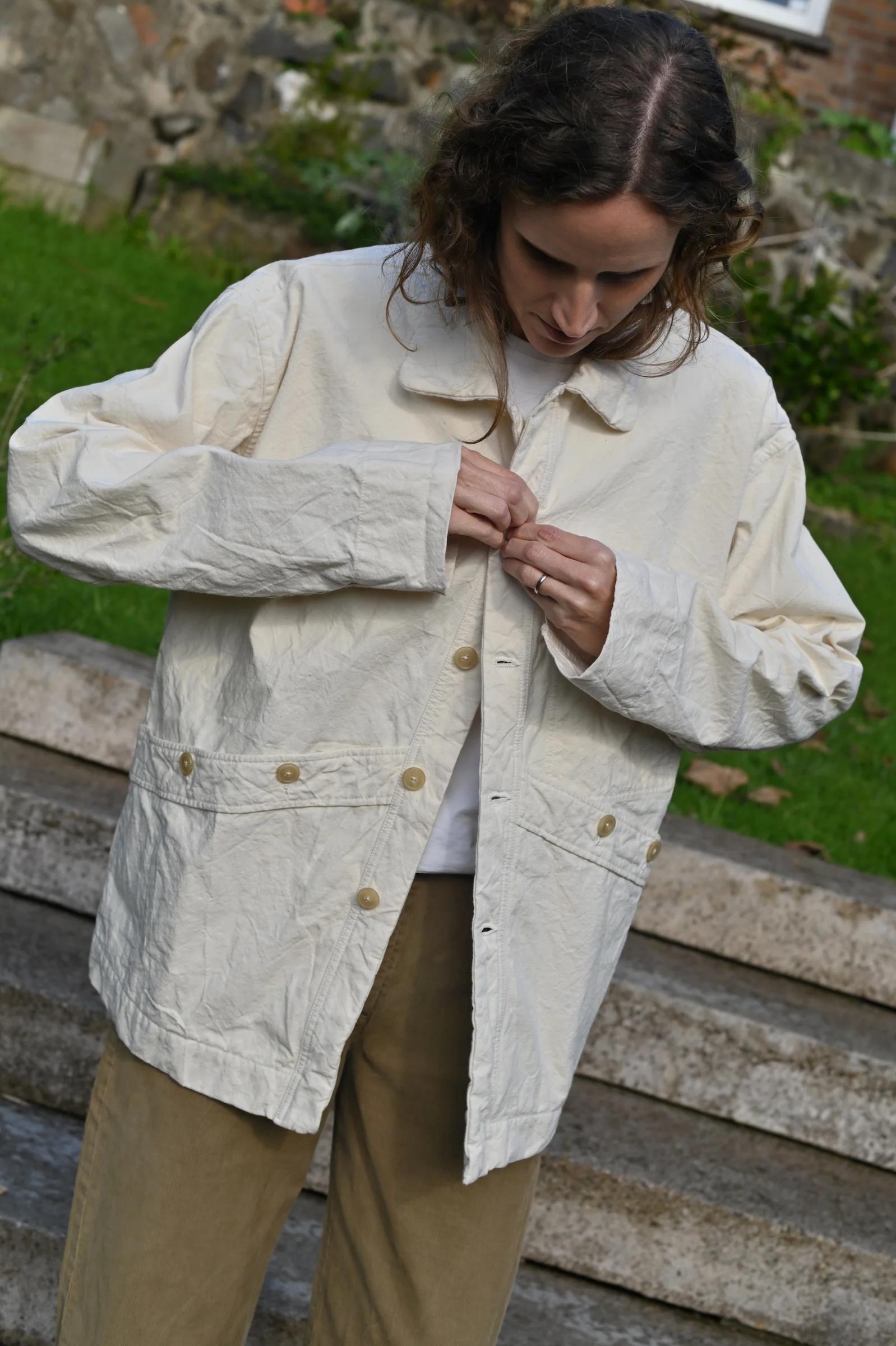 Curly-haired person buttoning light jacket outdoors. Tan pants, stone steps and wall with greenery in background.