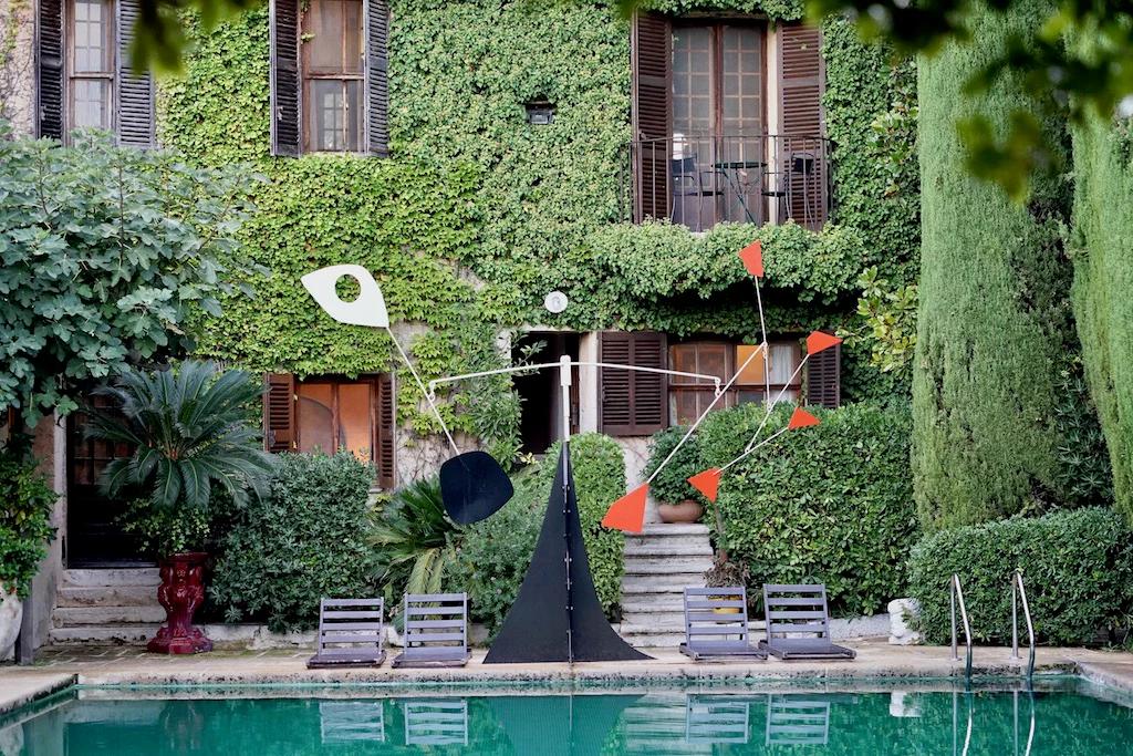 Courtyard with pool, loungers, and modern sculpture. Rustic building with shutters and balconies. Lush greenery surrounds.