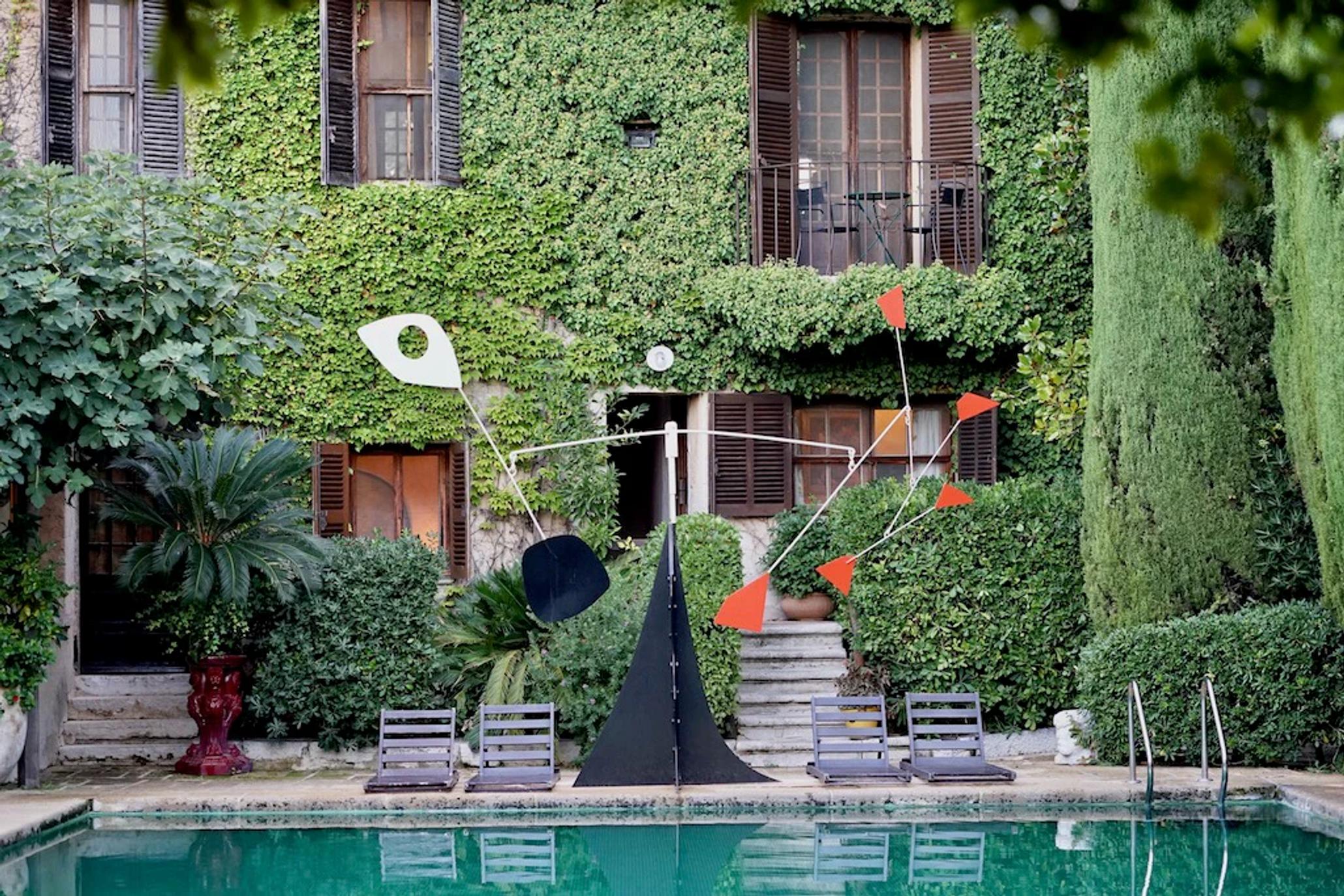 Courtyard with pool, loungers, and modern sculpture. Rustic building with shutters and balconies. Lush greenery surrounds.