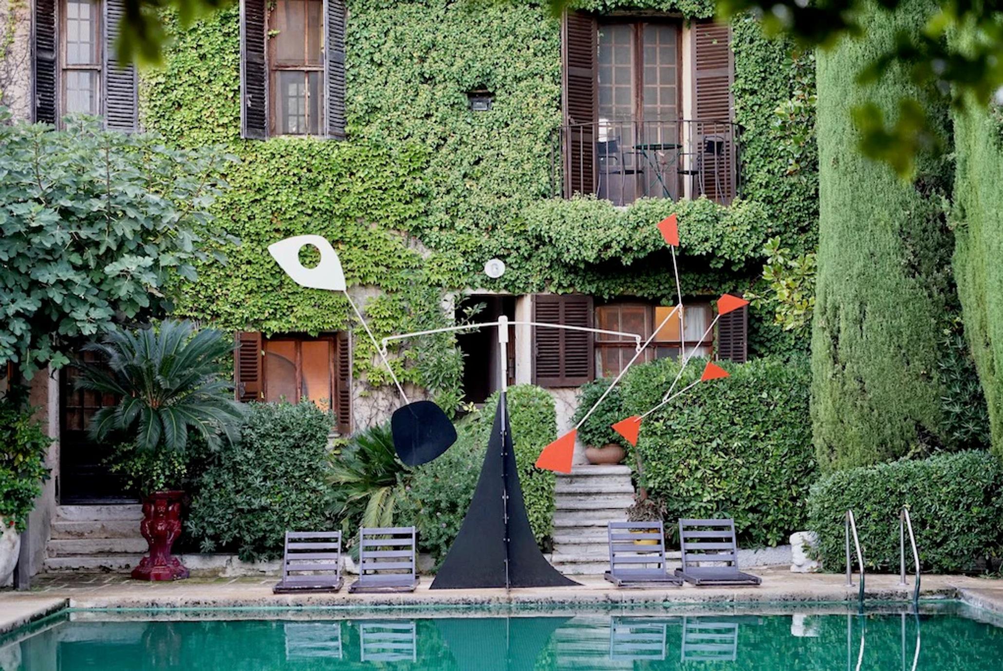 Courtyard with pool, loungers, and modern sculpture. Rustic building with shutters and balconies. Lush greenery surrounds.