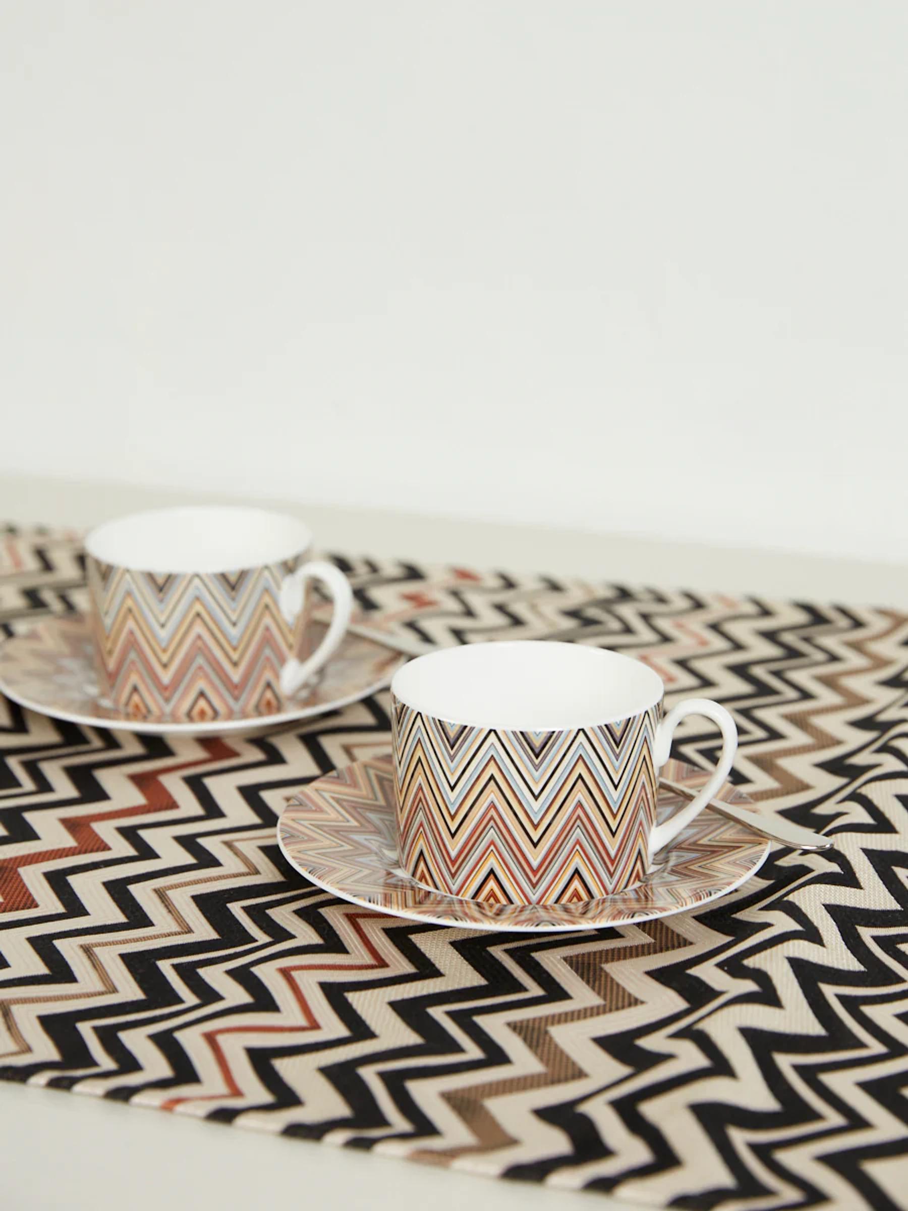 Two white teacups with zigzag design on brown, black, and beige zigzag patterned runner. White wall background.