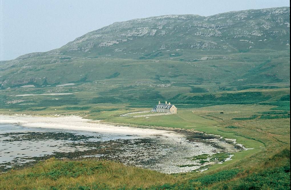 Stone cottage on Jura's grassy shore. Rocky coastline, hills, and mountain in background. Serene, remote setting.