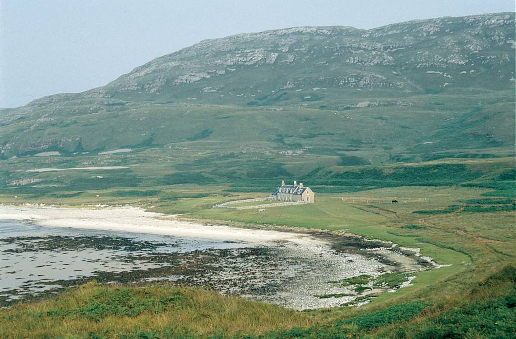 Stone cottage on Jura's grassy shore. Rocky coastline, hills, and mountain in background. Serene, remote setting.