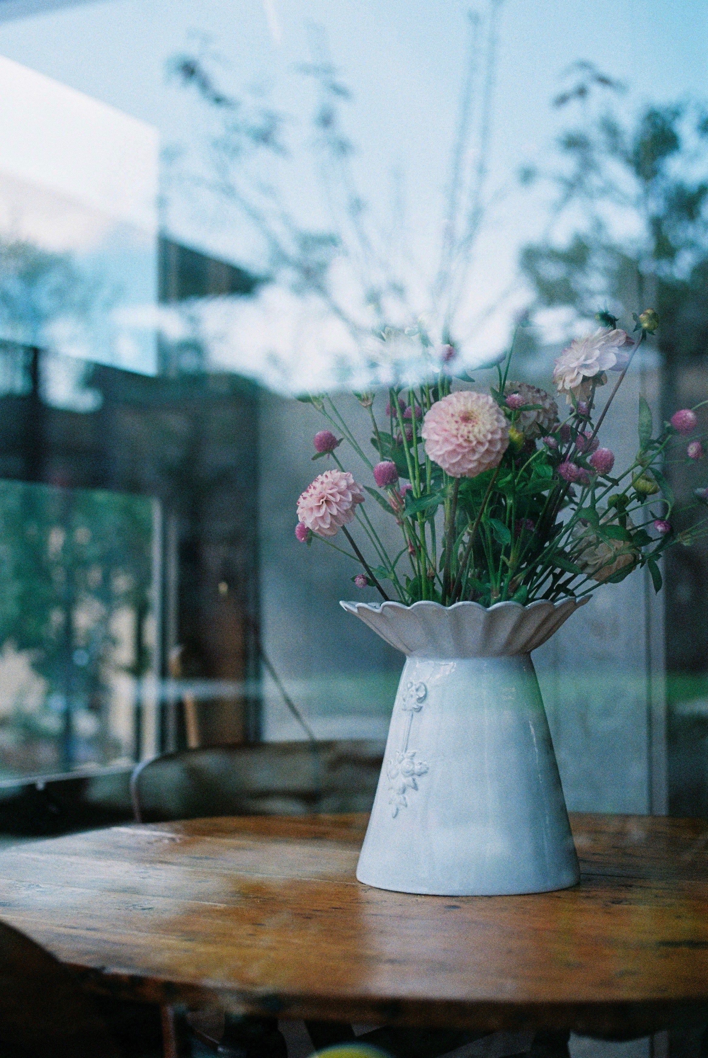 White vase with pink and white flowers on wooden table by window. Window reflects outdoor greenery and blue sky.