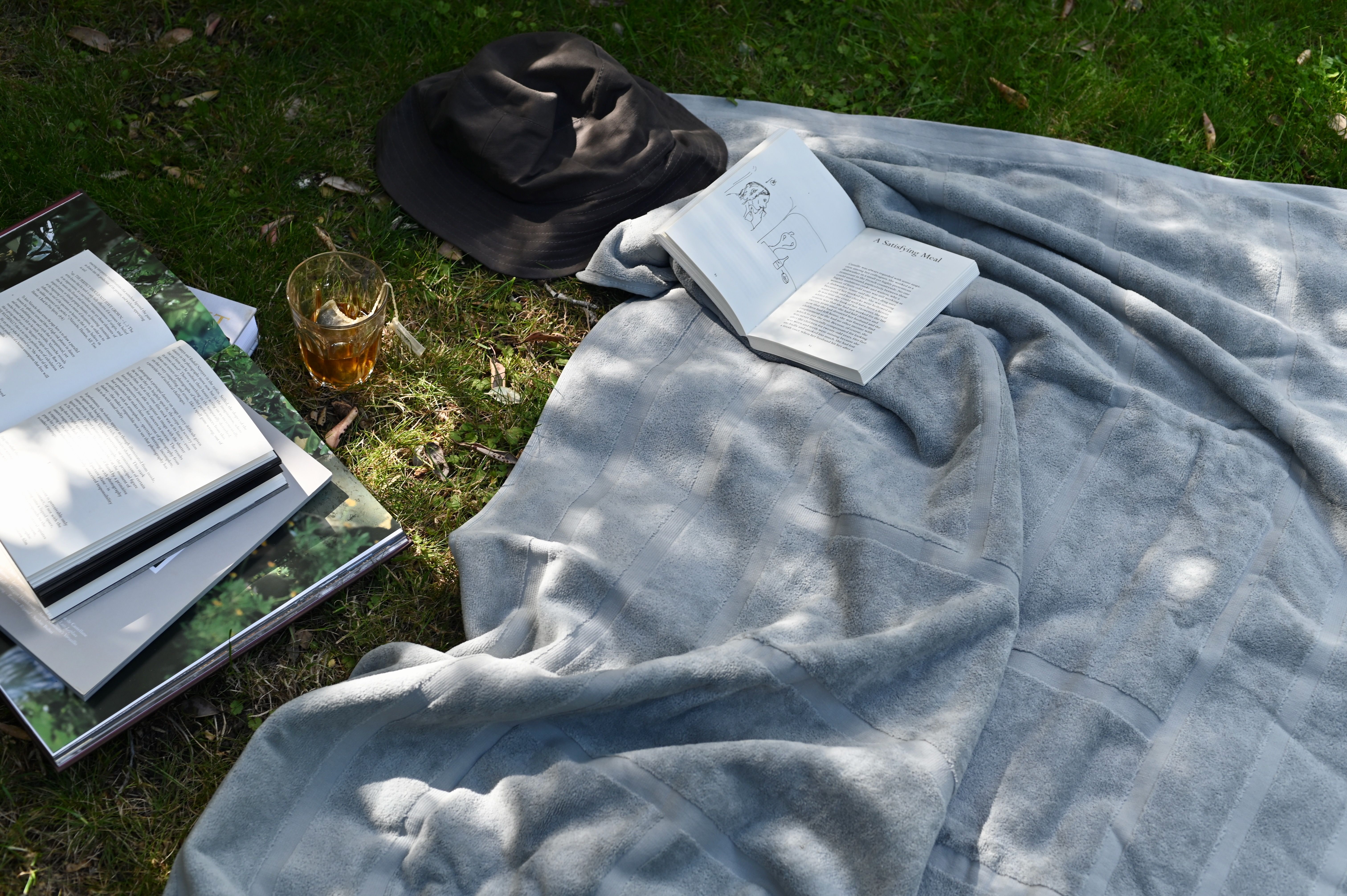 a soft towel lays on the grass next to a pile of books and sunhat 