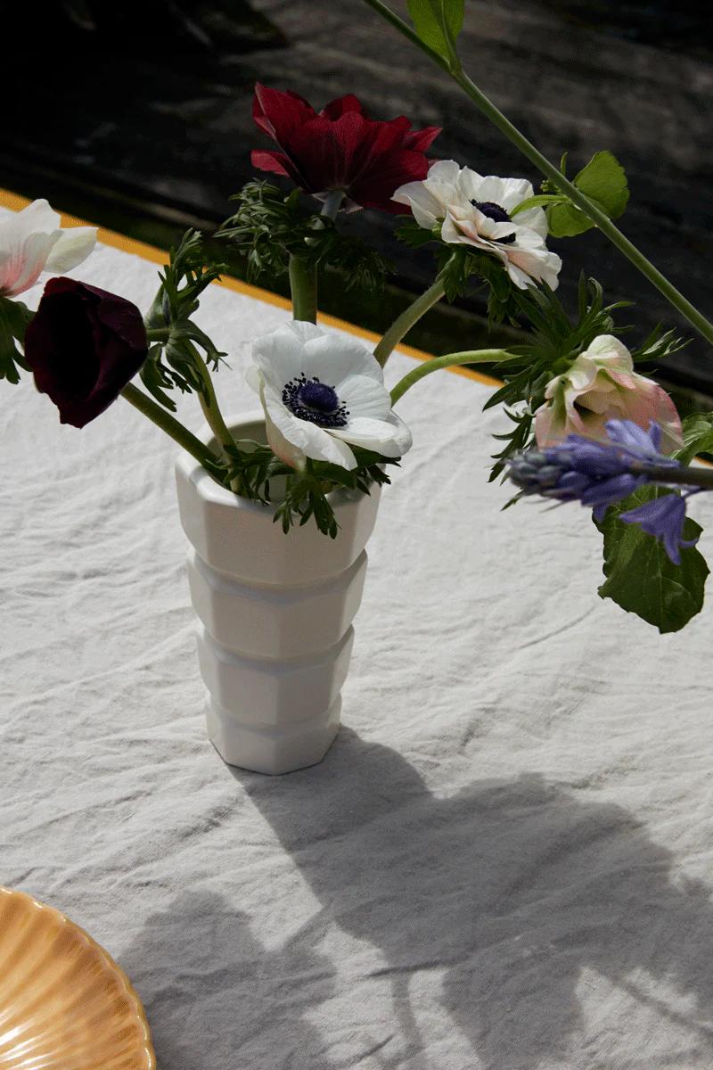 A white vase with maroon, white, and purple flowers sits on a table with a light tablecloth. Sunlight casts shadows, while part of a gold-rimmed plate enhances the dinner party scene.