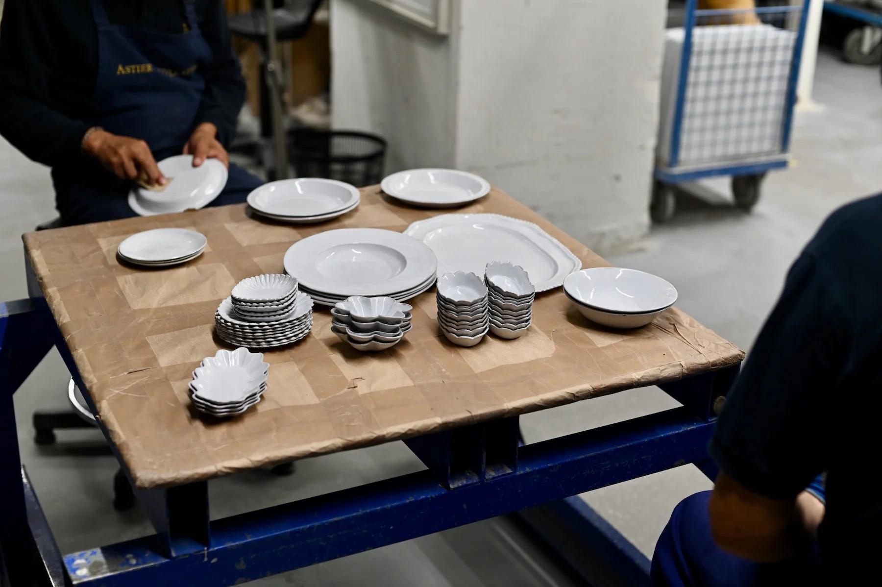 Two individuals organizing white ceramic plates and dishes on wooden table covered with brown paper. Shopping cart in background.