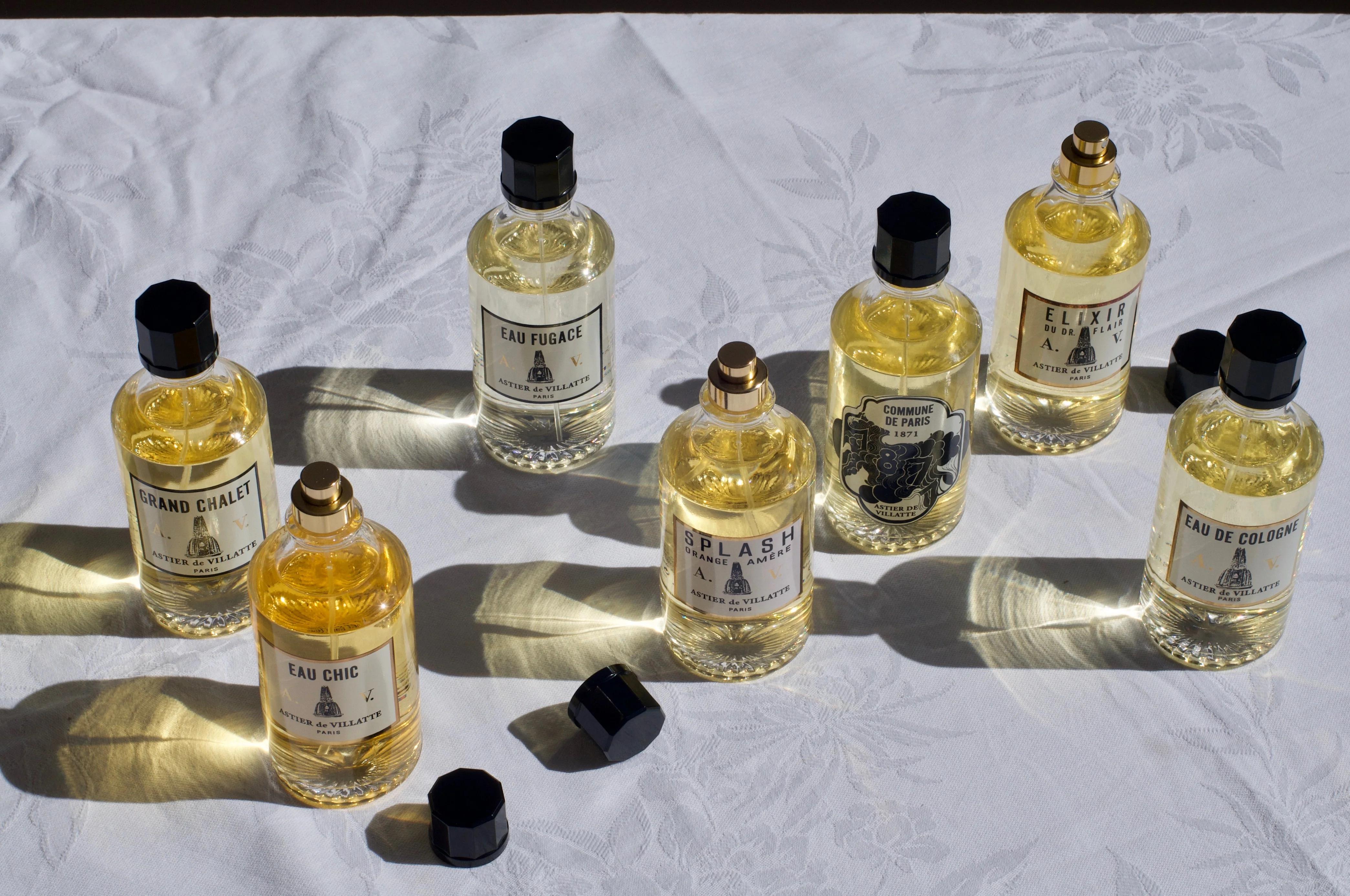 Seven Astier de Villatte perfume bottles with black caps on a white patterned tablecloth. Two caps are off, with sunlight casting stunning shadows and reflections across the scene.