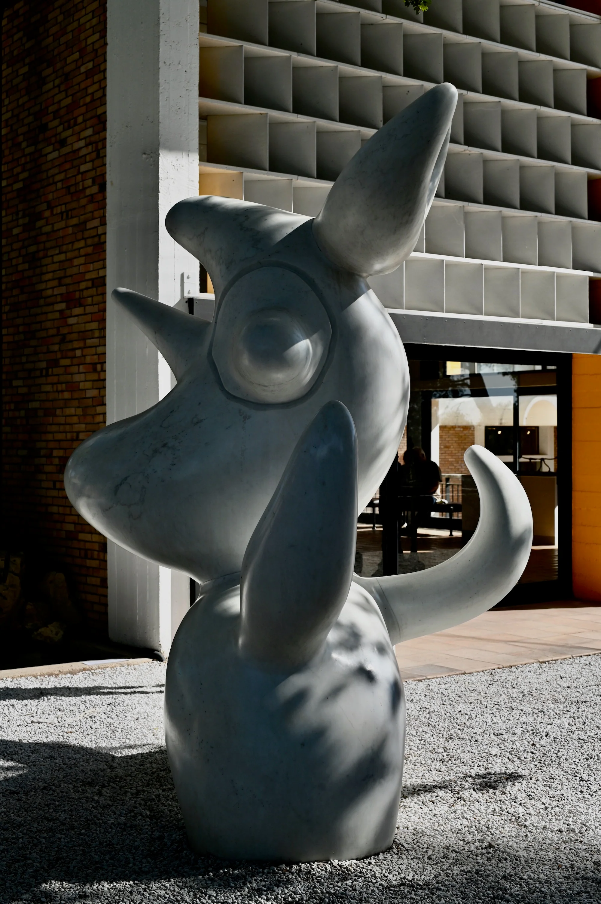 Abstract animal sculpture on gravel. Exaggerated features. Modern building with grid facade in background.