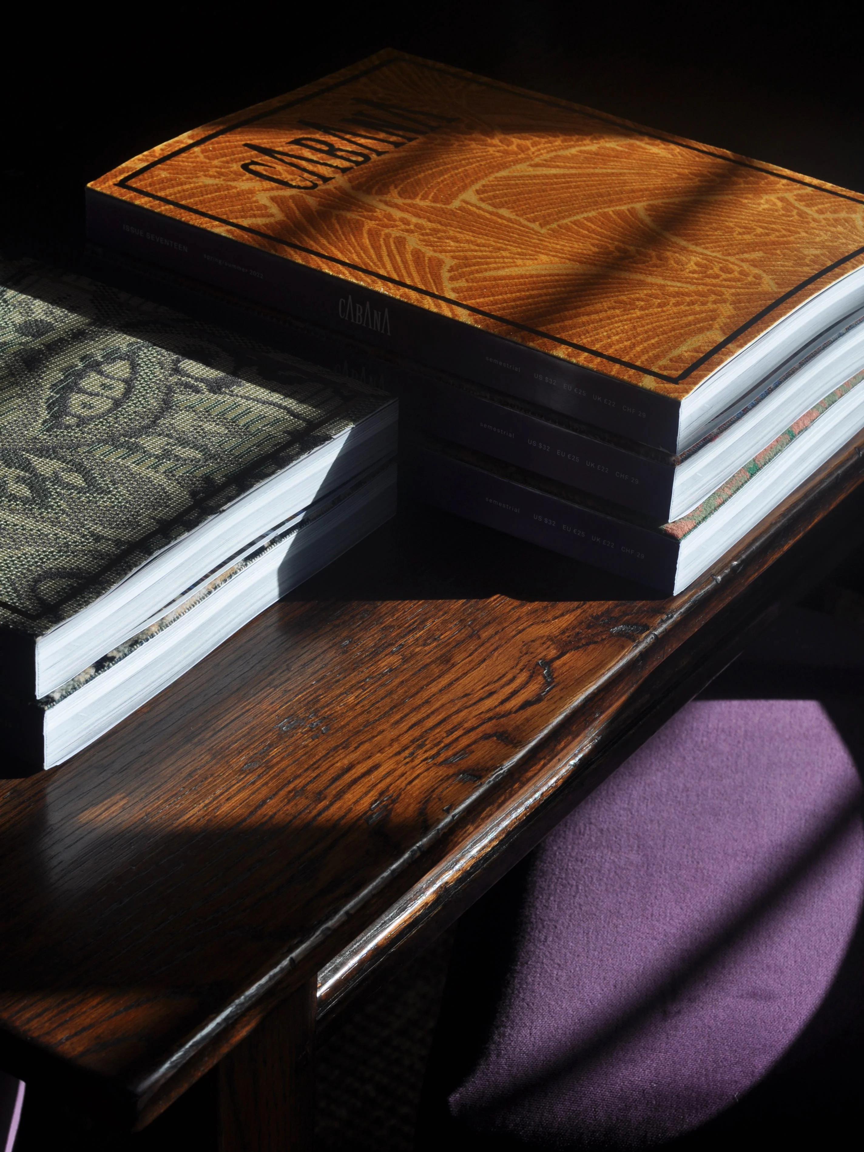 Stack of textured books on sunlit wooden table. Purple chair partially visible. Shadows creating contrast.