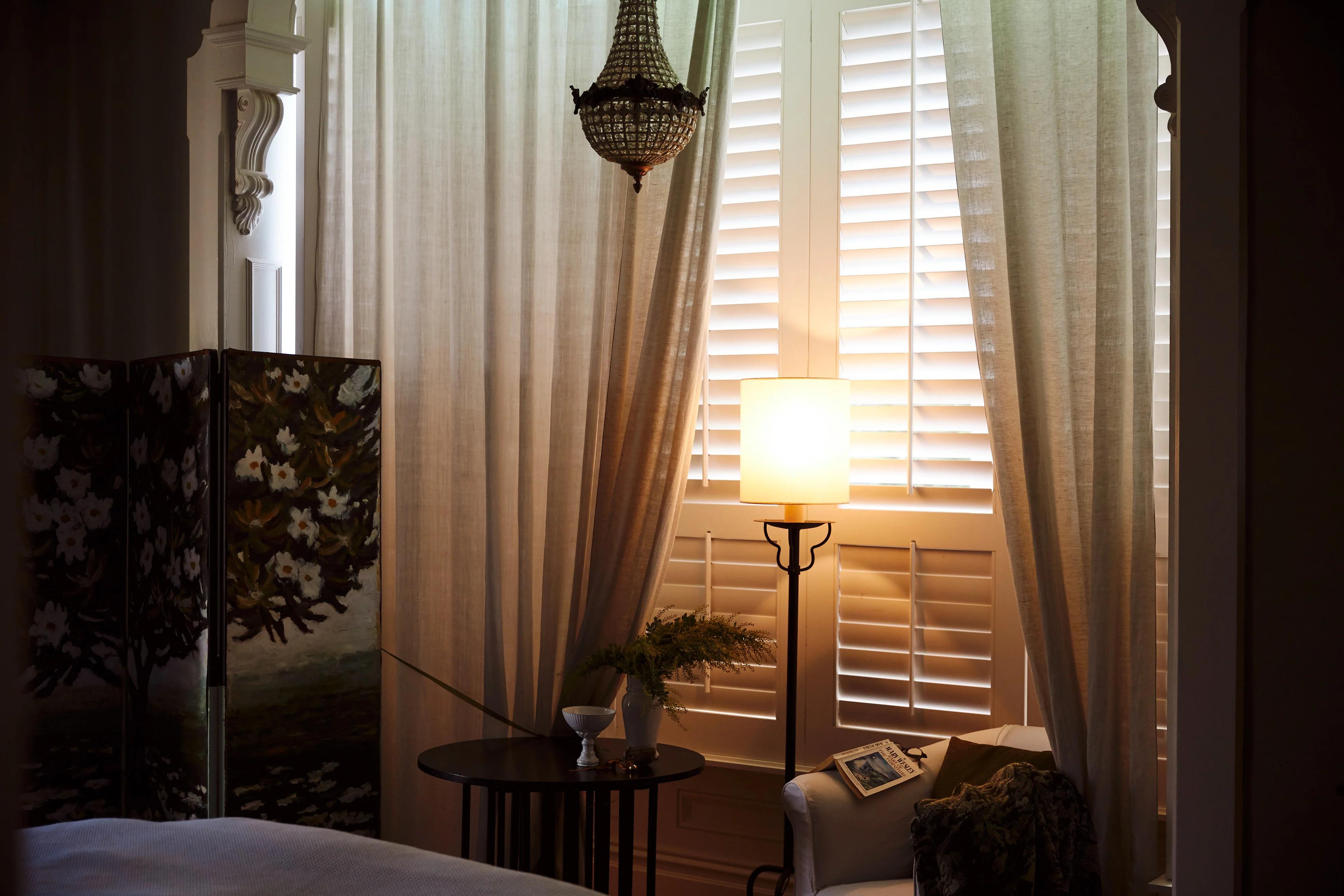 Cozy room with chandelier, curtains, shutters. Folding screen in corner. Side table with Balthus Lamp, books, vase.