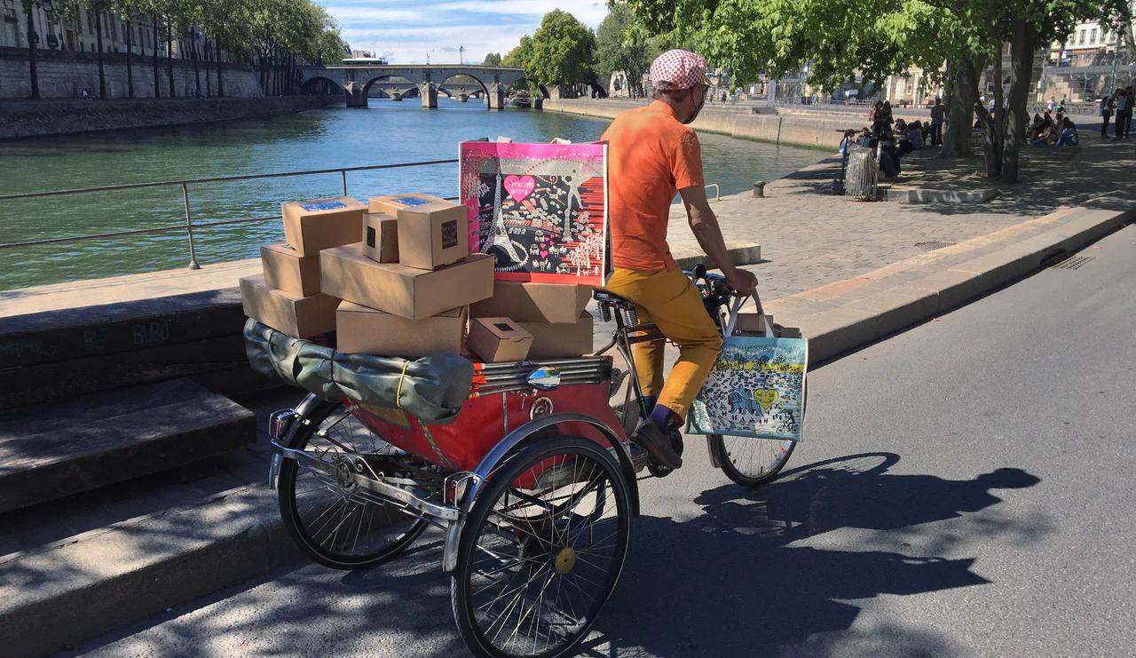 Person on red tricycle carrying boxes along Parisian riverside. Wearing bright clothes. Trees and bridge visible in background.