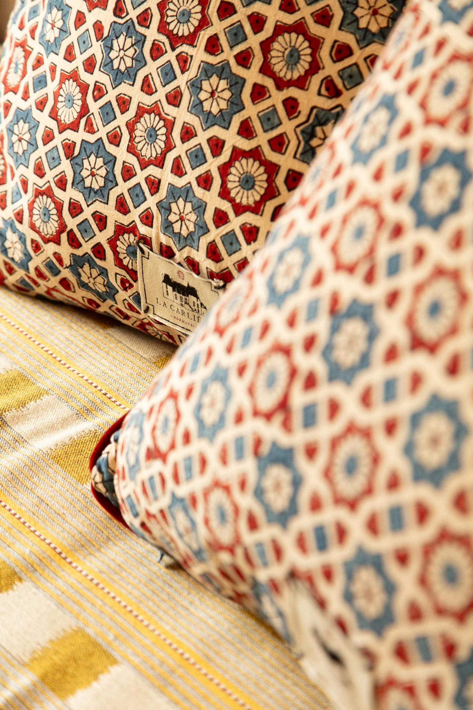 Close-up of geometric and floral pillows in red, blue, beige. Yellow striped fabric underneath. Small label visible.