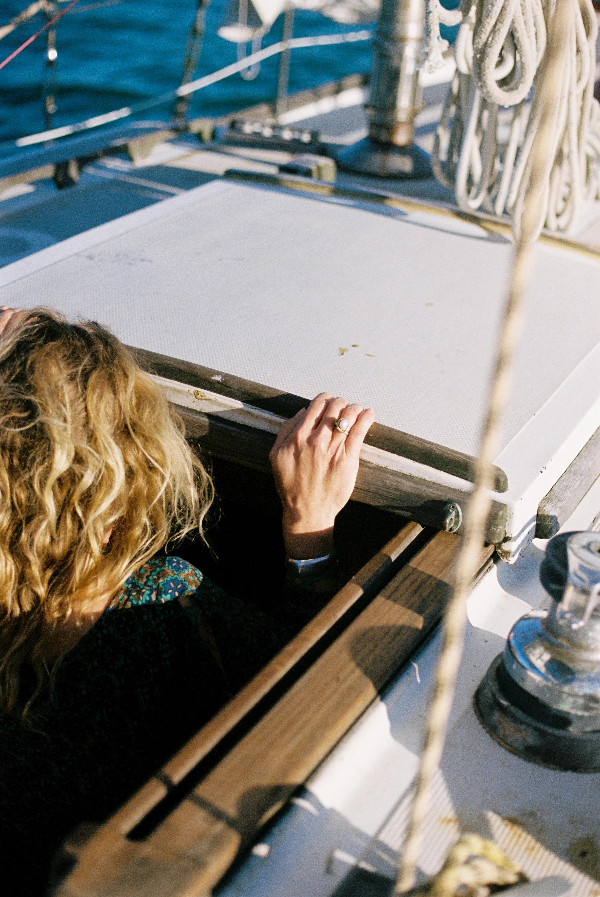 Person with curly blonde hair emerging from boat hatch. Hand on hatch edge, ropes visible on deck.