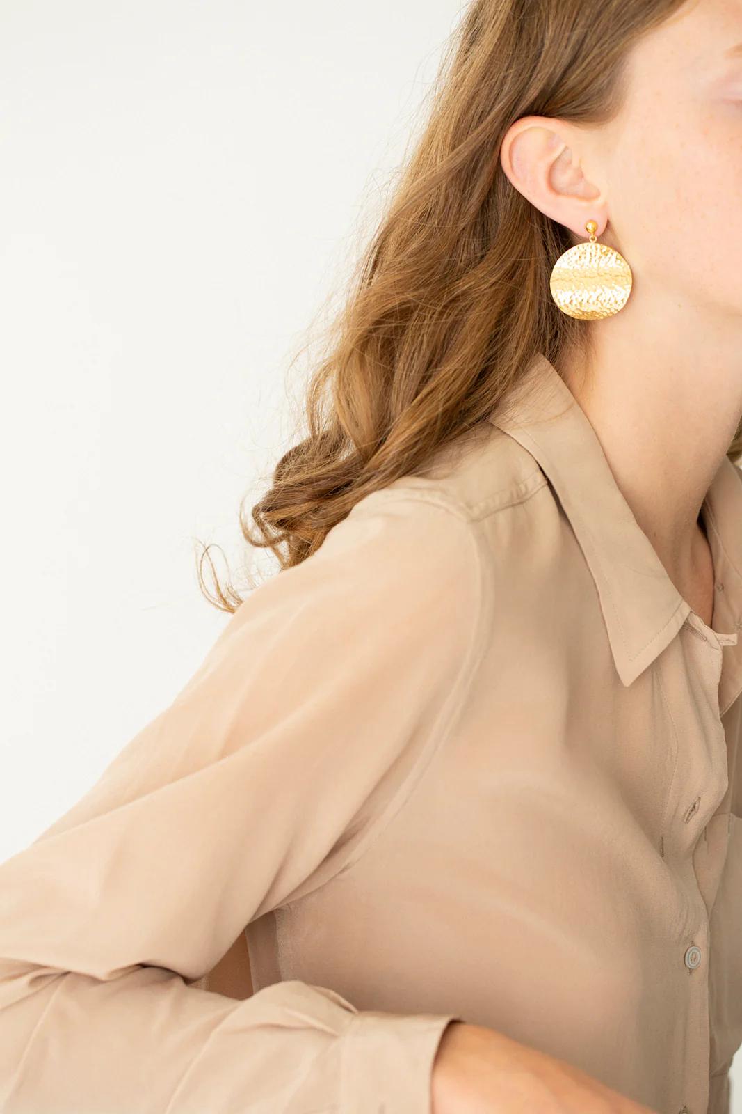 Close-up of long-haired person in sheer beige shirt. Large, textured gold earrings. Minimalist aesthetic with Charlotte Penman jewellery touch.